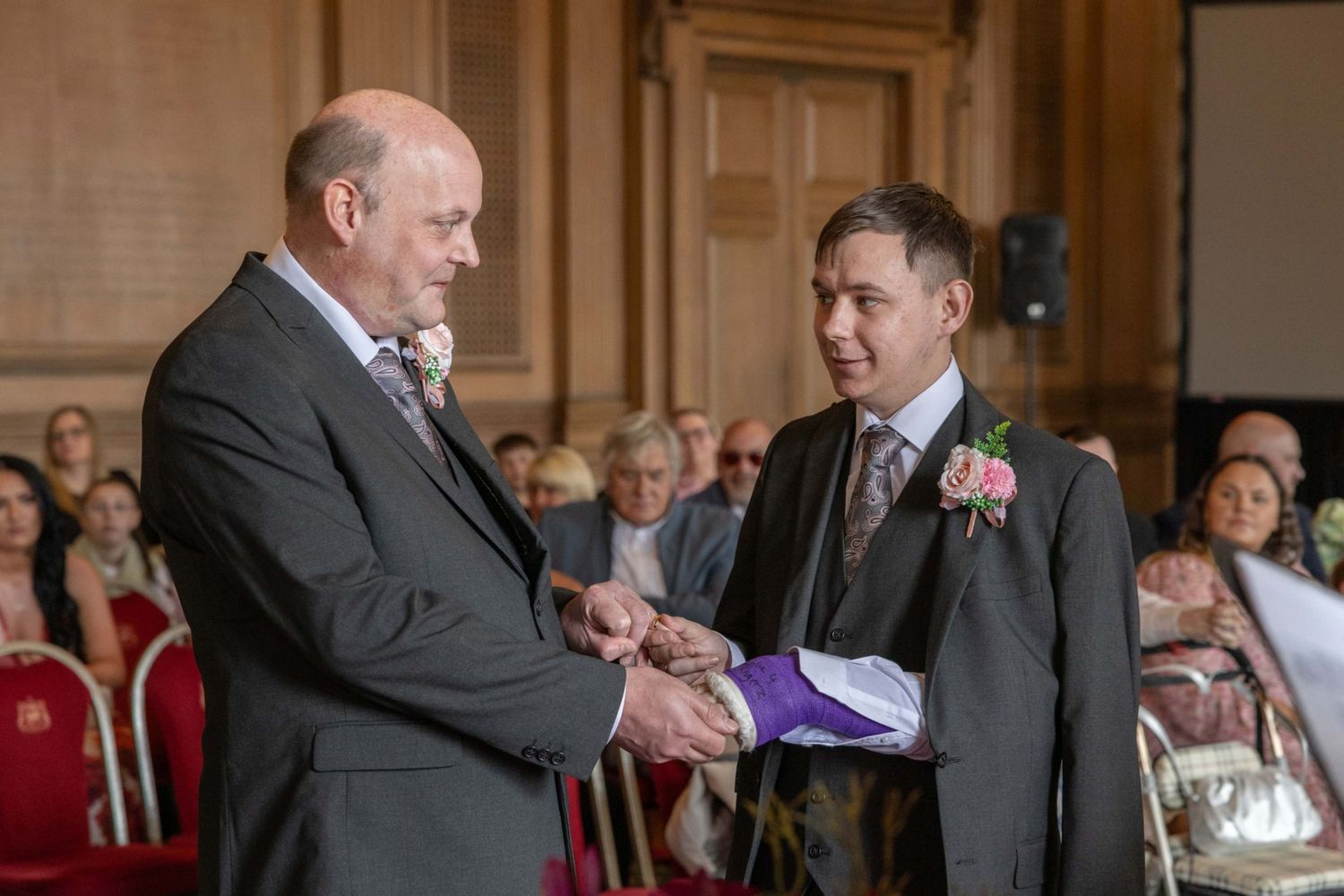 Two grooms exchange rings at a wedding ceremony in an elegant venue with guests seated in the background.