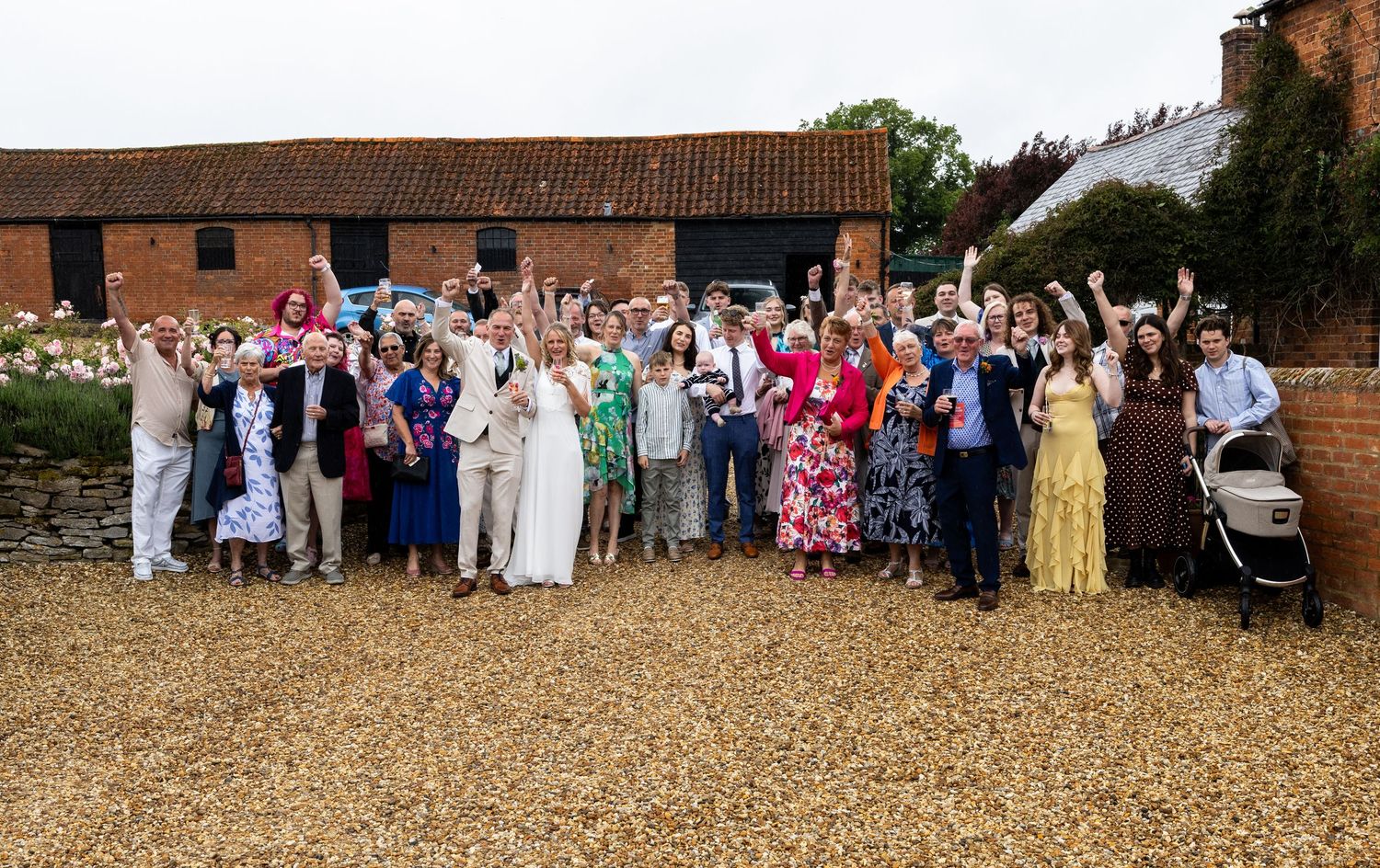 Large wedding group photo outdoors at rustic barn venue, guests cheering with bride and groom in center.