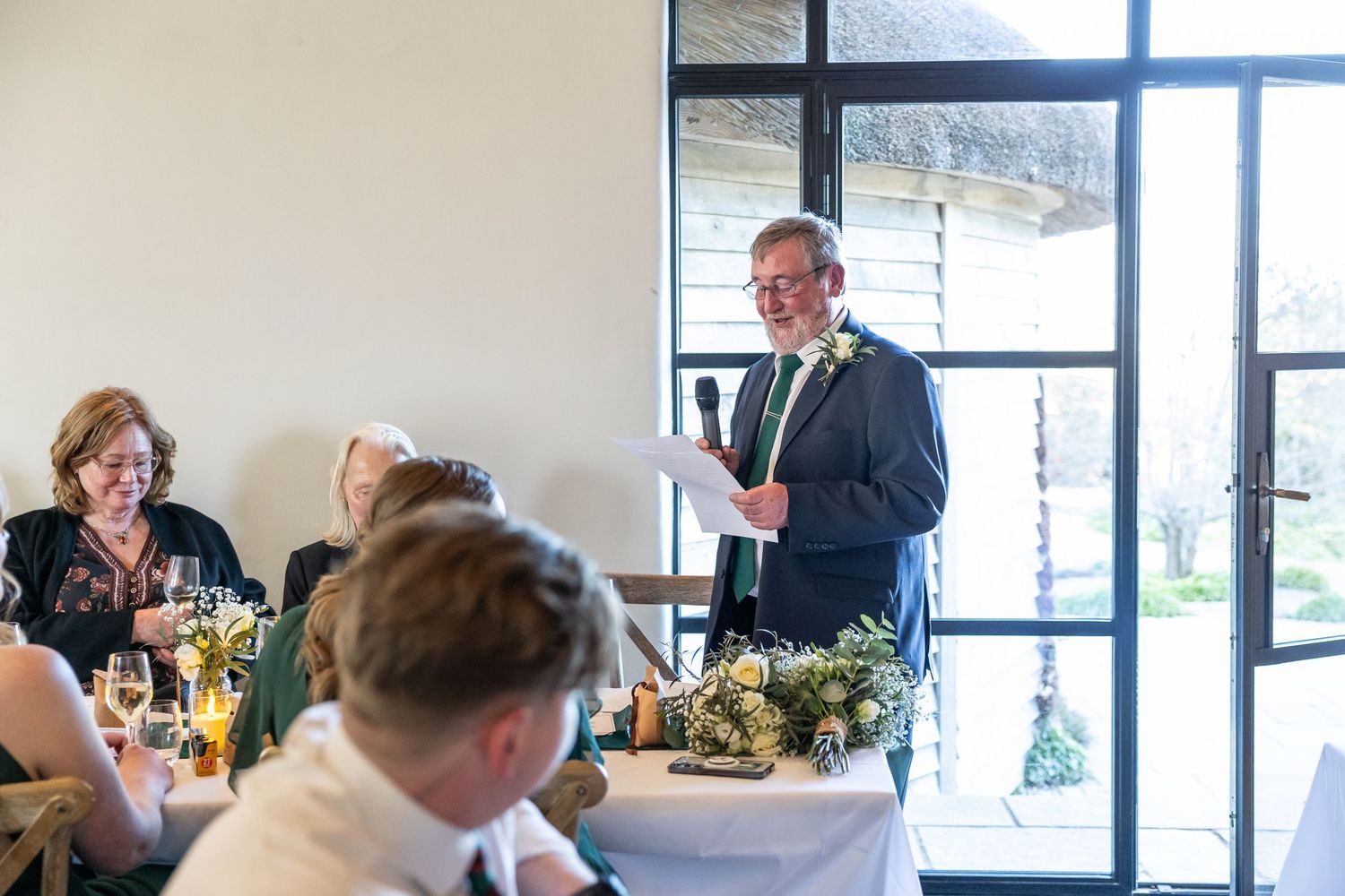 Man in suit giving a speech at a wedding reception, holding papers, smiling at guests seated at decorated tables.