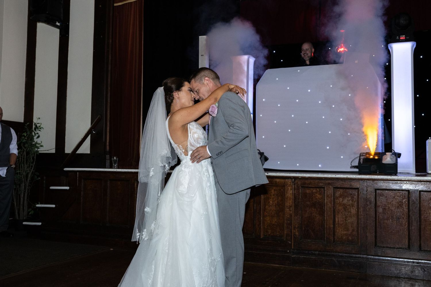 Newlyweds share romantic first dance kiss with smoke effects and DJ backdrop at wedding reception.