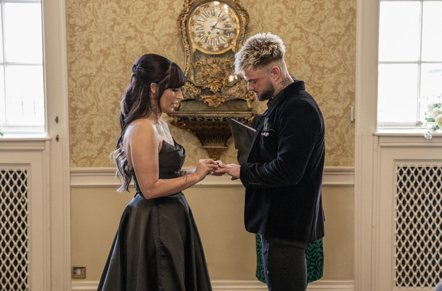 Couple exchanging rings in elegant wedding ceremony indoors, bride in black gown, groom in dark hoodie.