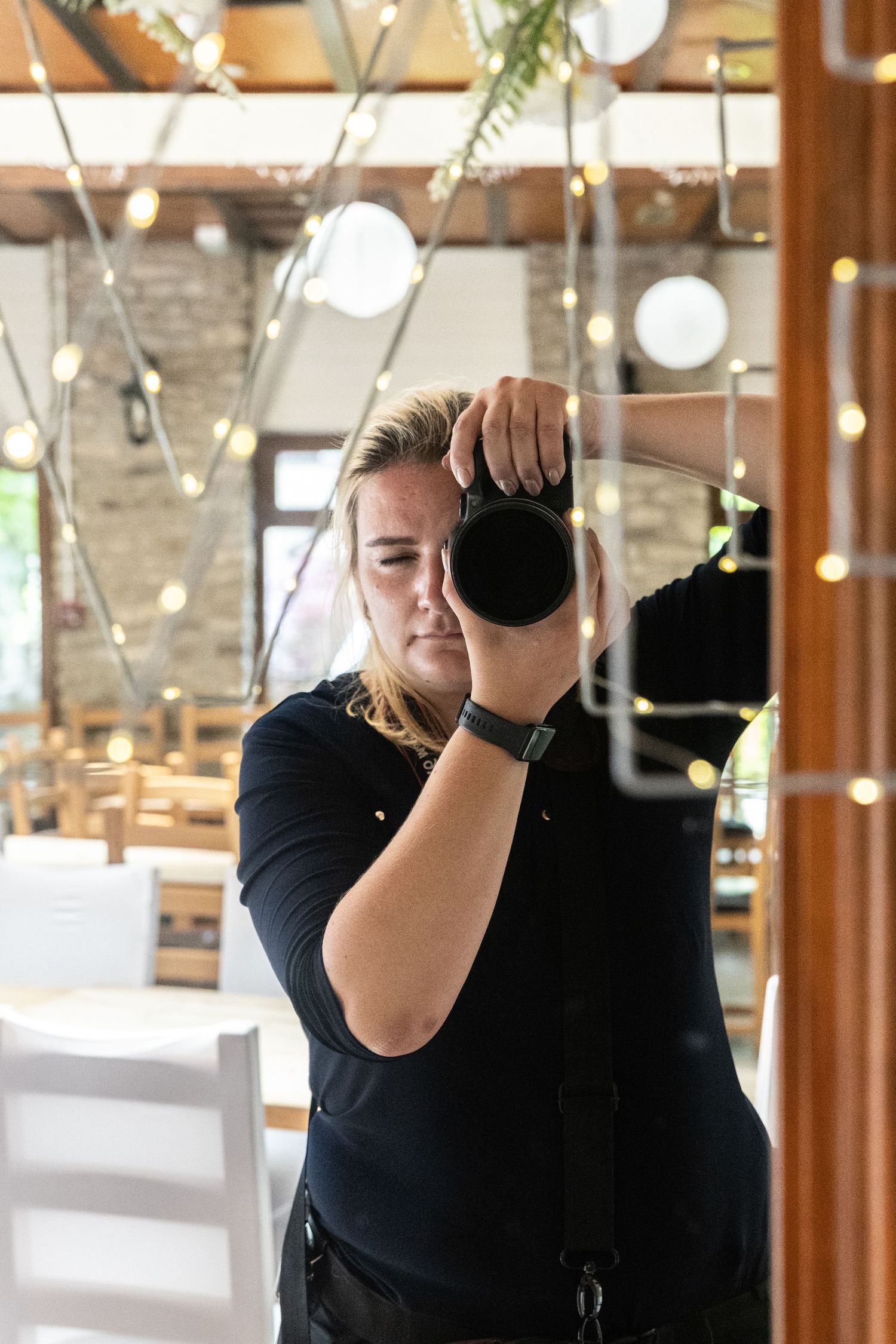 Photographer in black shirt adjusts camera settings while working in a bright indoor venue with wooden decor.