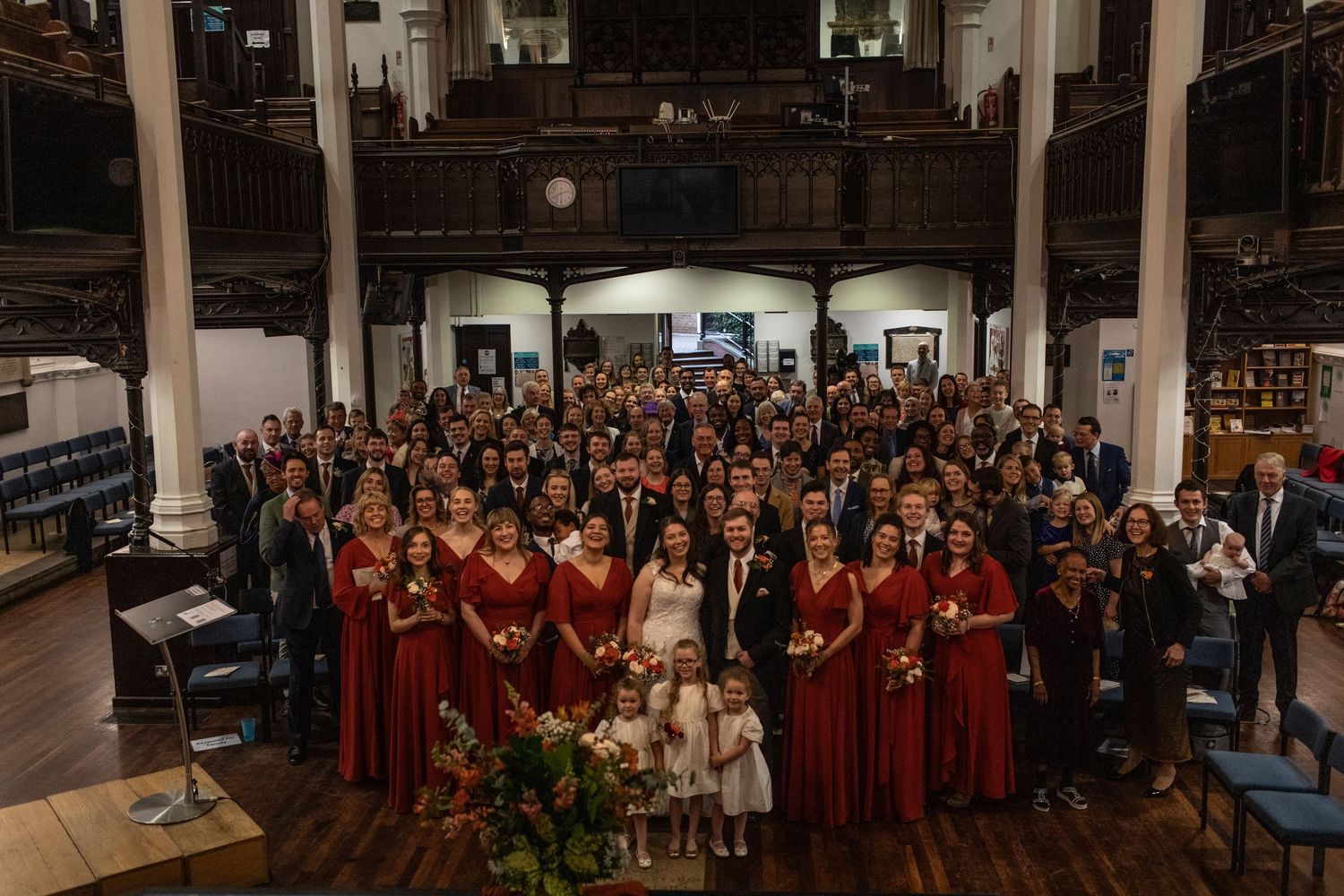 Large wedding group photo with bridesmaids in red dresses and guests gathered in an elegant historic venue.