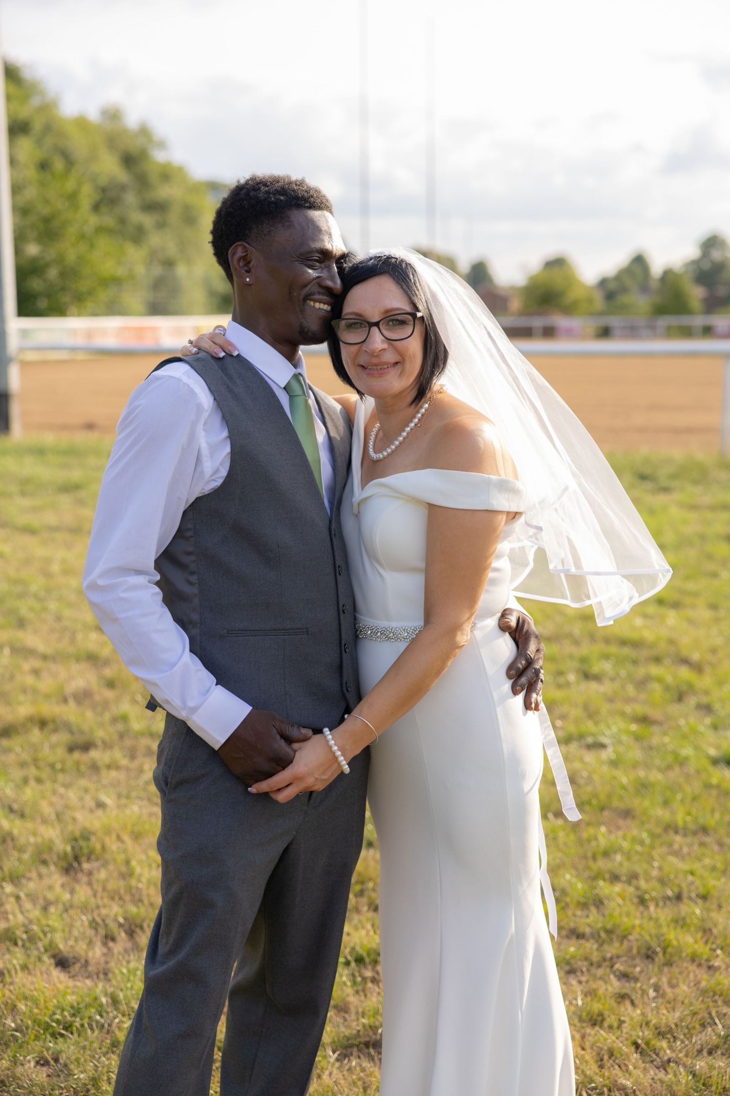 Interracial wedding couple posing outdoors on grass, groom in gray suit with green tie, bride in white gown and veil.