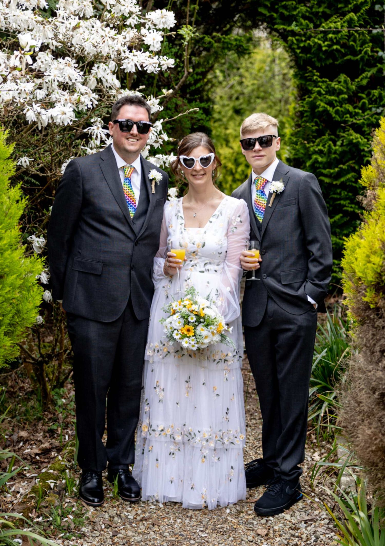 Bride in floral dress with bouquet poses with two groomsmen in gray suits and rainbow ties wearing sunglasses.