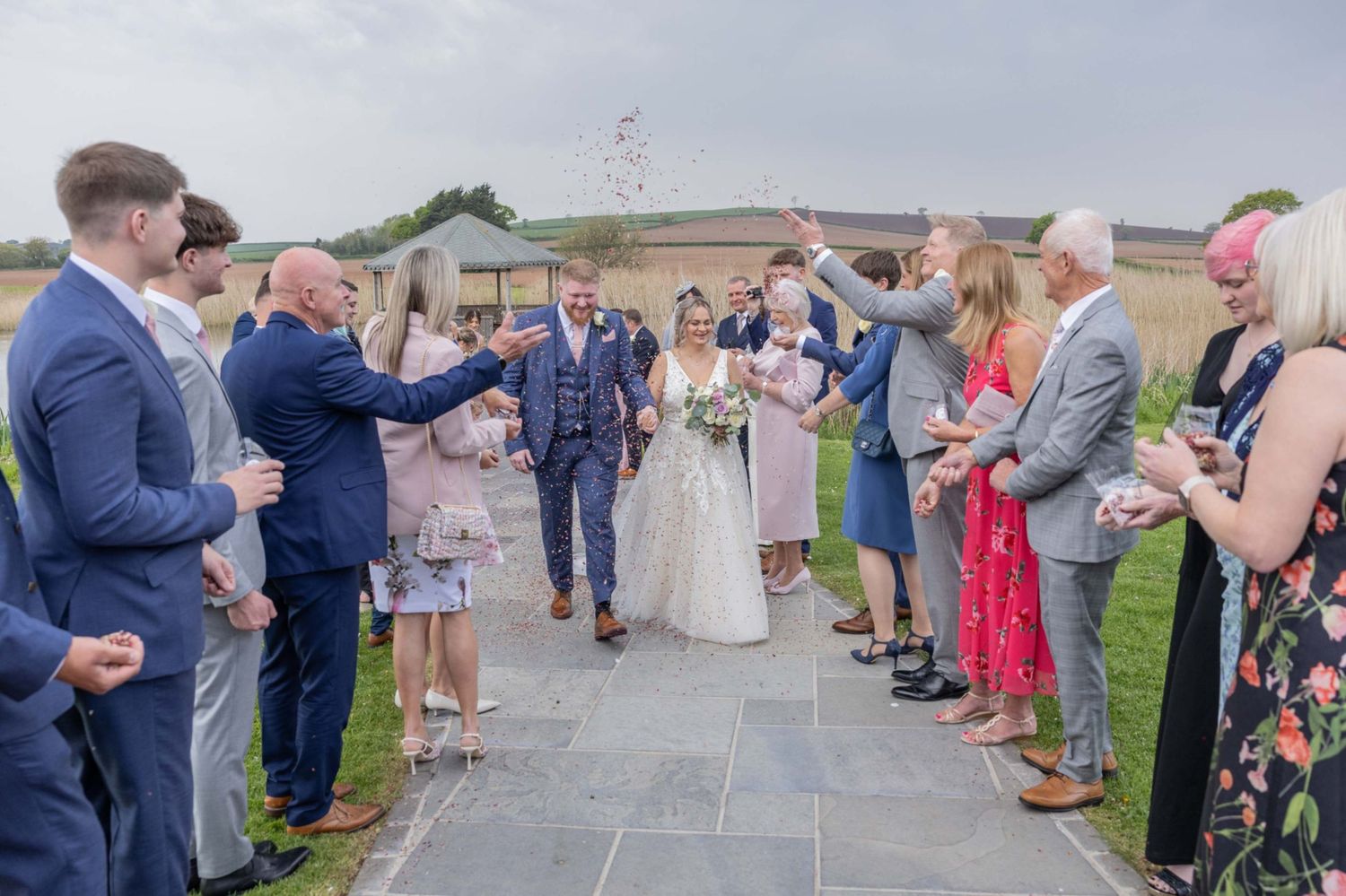 Bride and groom walk through cheering guests throwing confetti outside a rustic wedding venue on a cloudy day.
