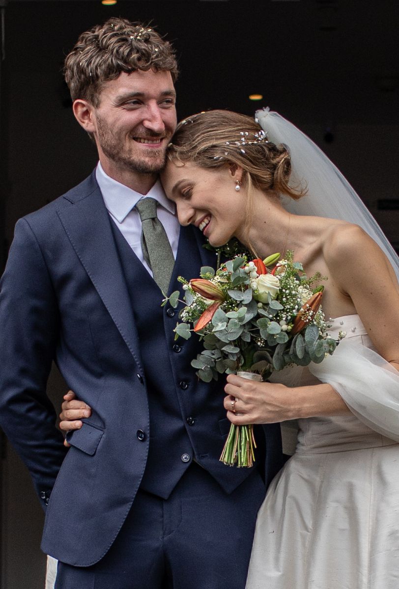 A smiling groom in a navy suit and bride in a white gown with veil holding a bouquet laugh together on their wedding day.