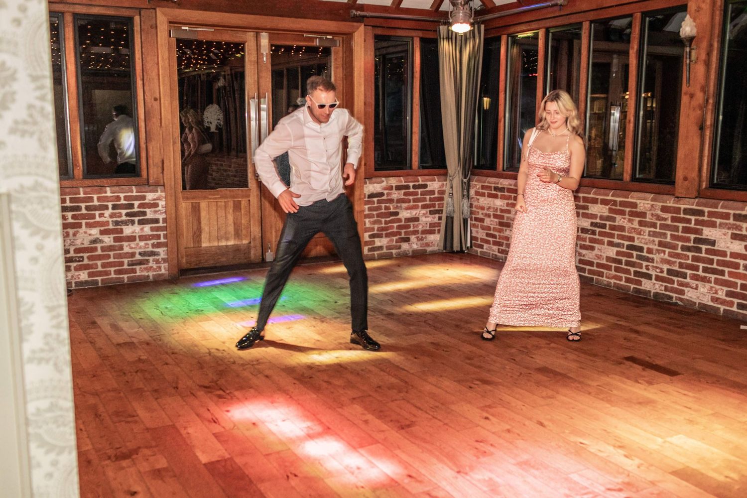 Couple dancing on a hardwood floor at a wedding reception in a rustic brick-walled venue with colorful lighting.