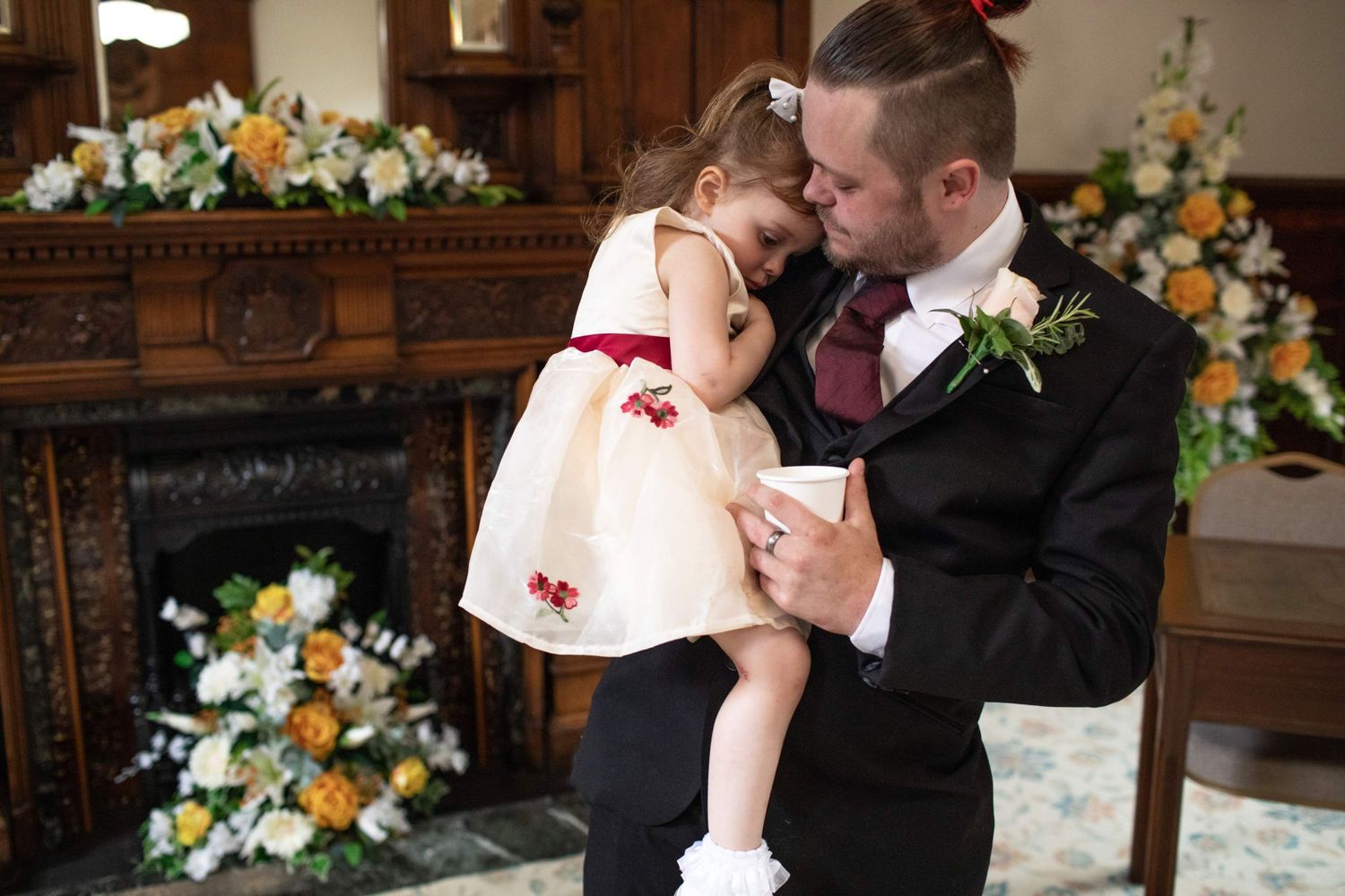 Groom in black suit tenderly holding young flower girl in cream dress at wedding ceremony.