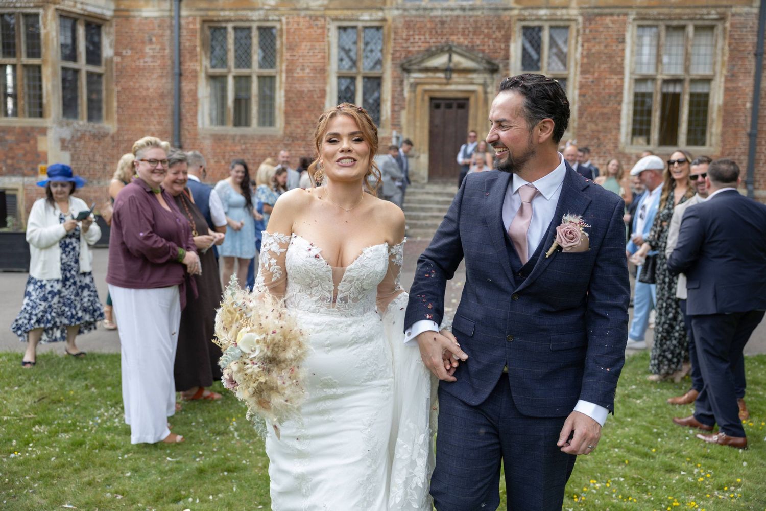 Bride and groom walk hand in hand outside a grand brick manor, surrounded by wedding guests celebrating their special day.