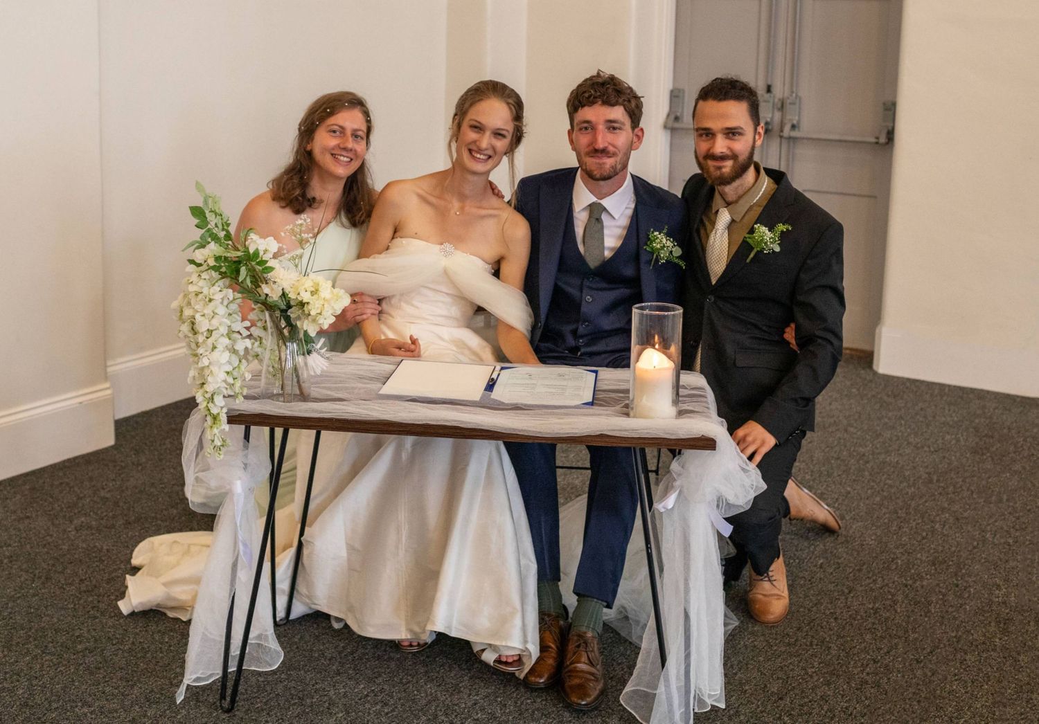 Bride and groom sit at a decorated table with two guests, celebrating their wedding reception indoors.