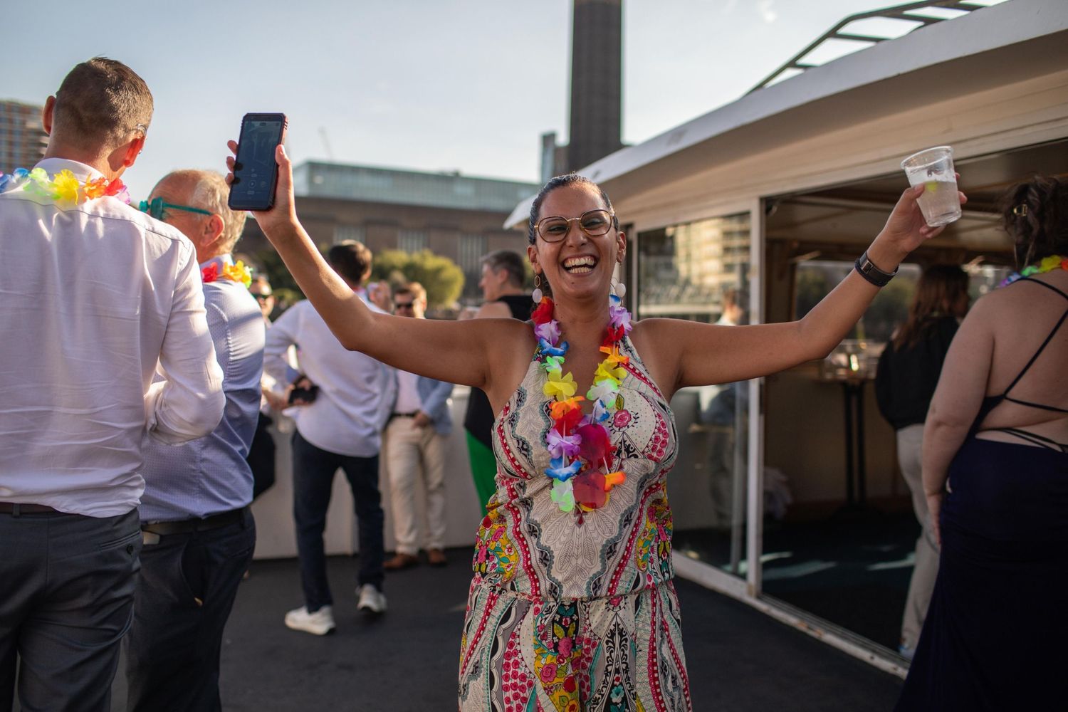 Woman in floral dress and lei celebrates outdoors holding phone at a colorful summer party event.