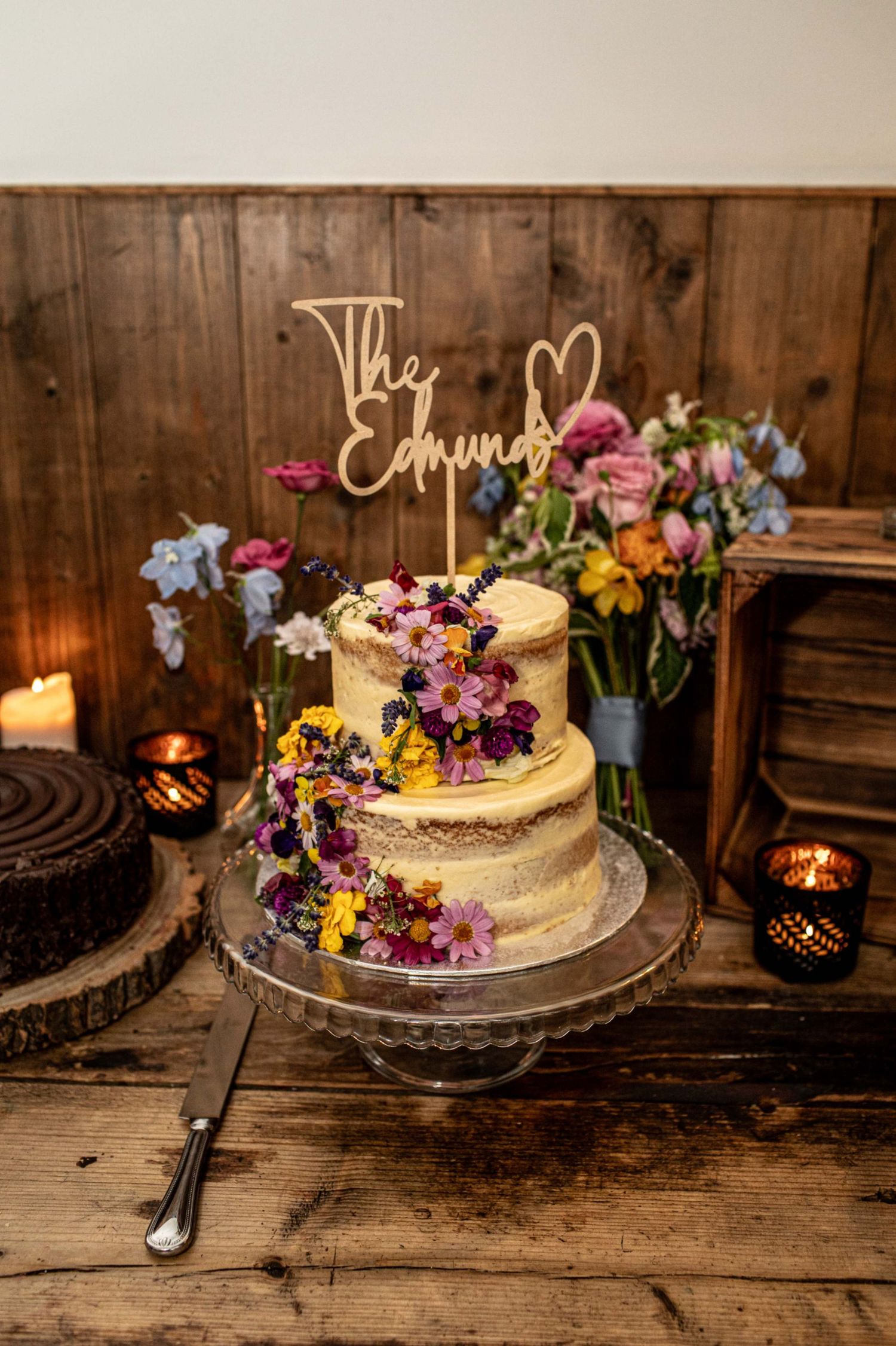 Two-tier semi-naked wedding cake decorated with colorful fresh flowers and a wooden The Edmunds cake topper on a glass stand.
