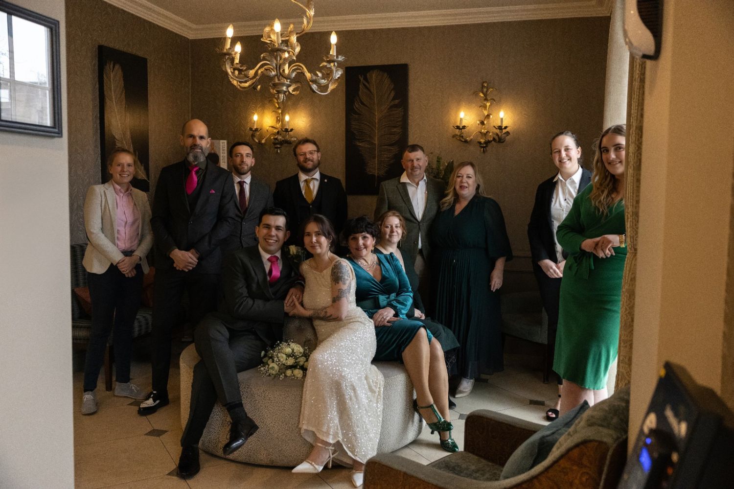 Wedding group photo with bride and groom seated, surrounded by family and friends in an elegant room with chandelier.