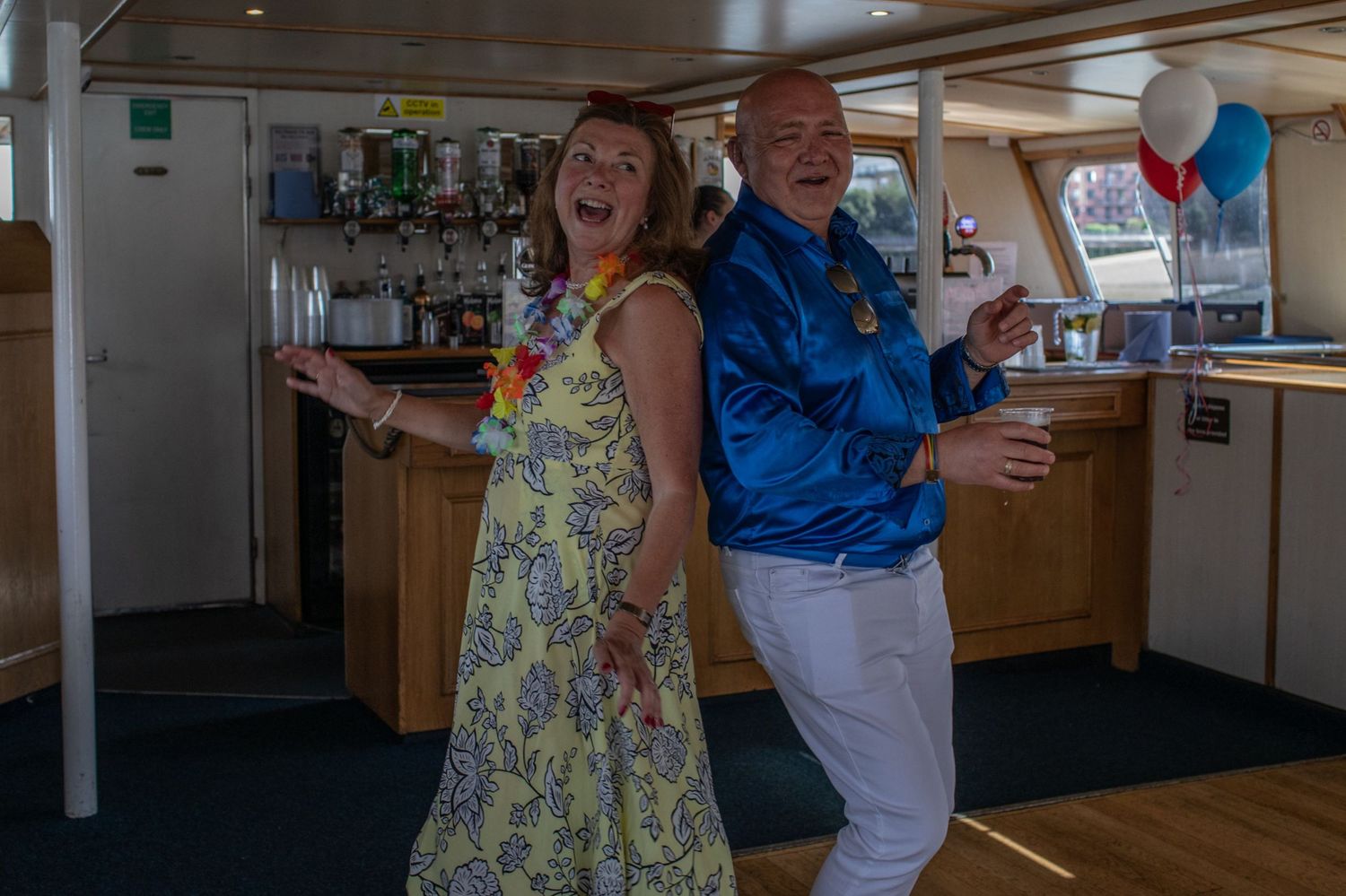 Joyful couple dancing on a cruise ship deck, woman in floral dress with lei, man in blue jacket, balloons in background.