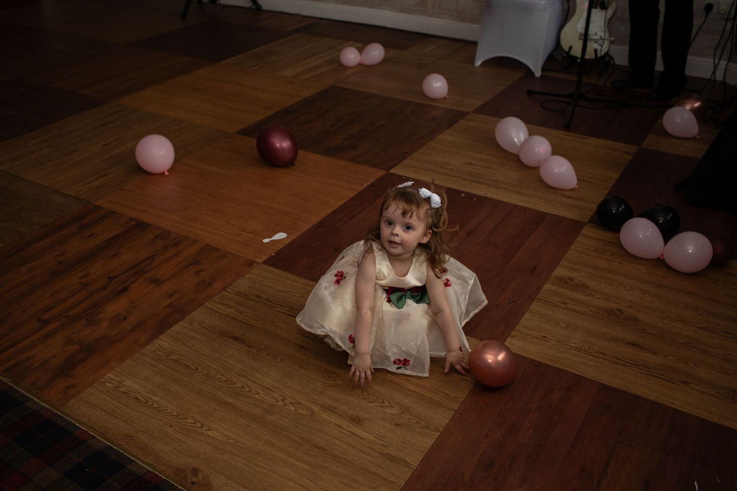 Young girl in white dress sitting on foam floor tiles surrounded by pink balloons at a birthday party.