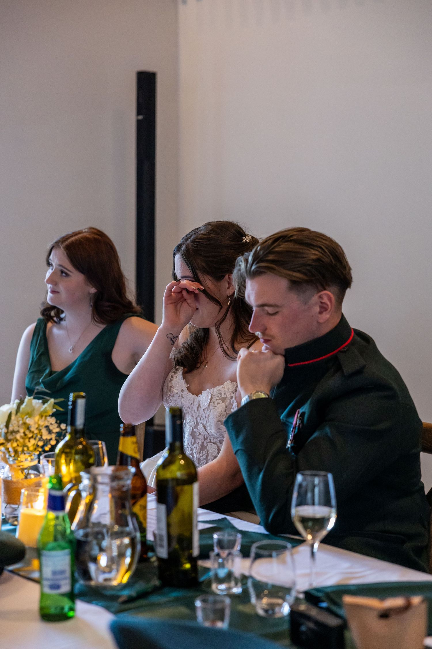 Bride and groom seated at wedding reception table with wine bottles, emotional moment during speeches.