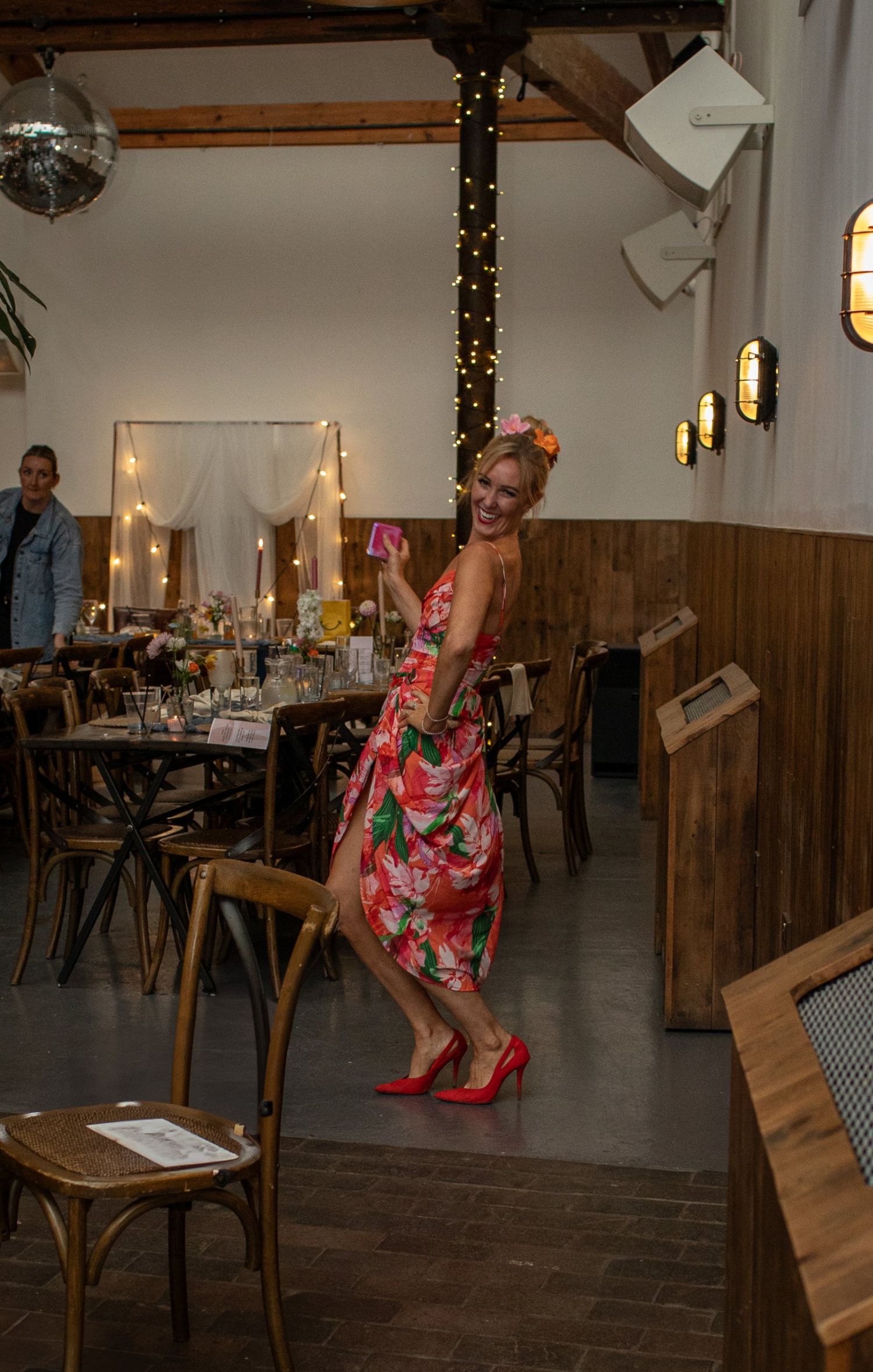 Woman in floral dress and red heels dancing joyfully at a rustic wedding venue with string lights.