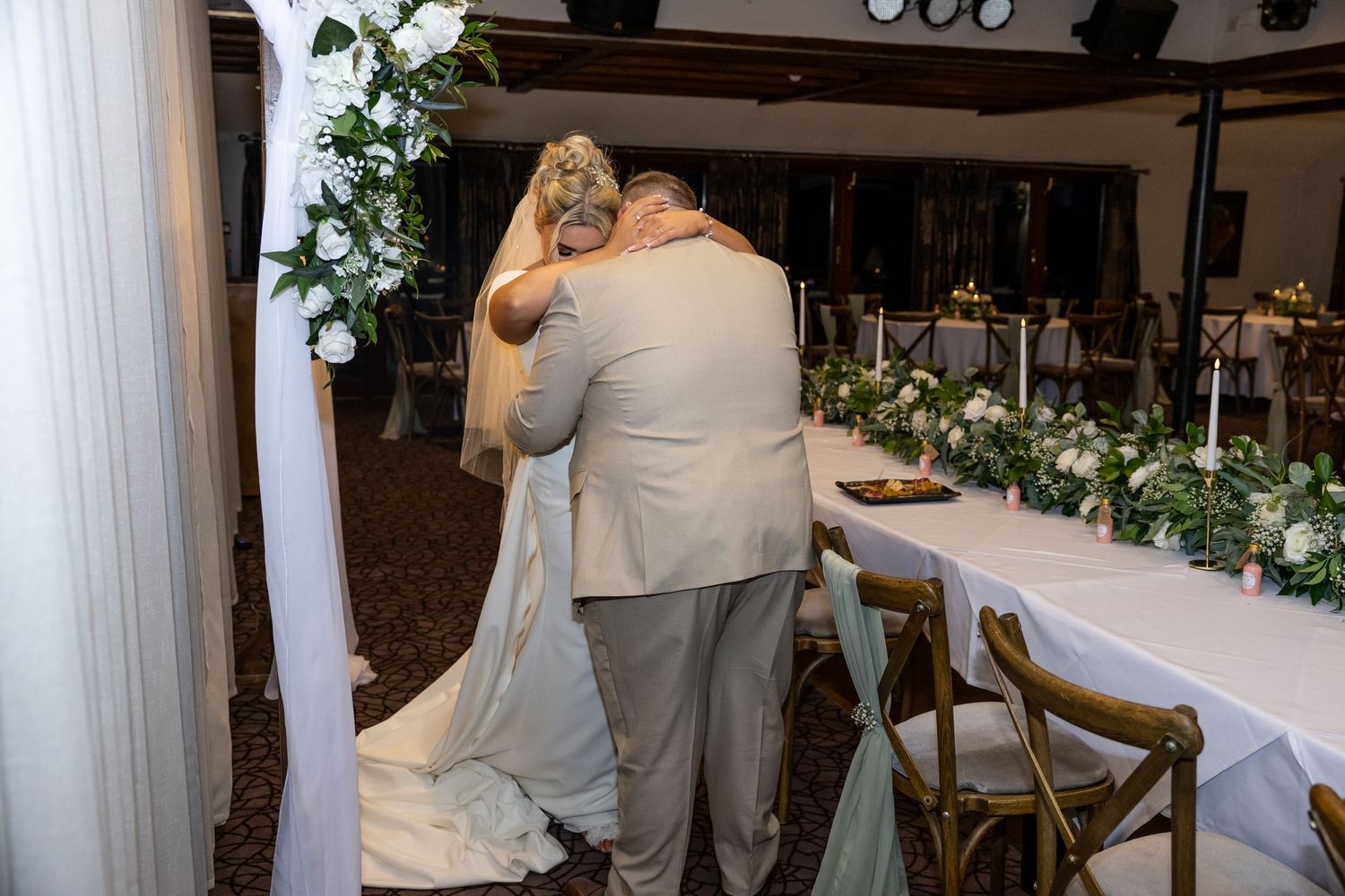 Bride and groom share emotional first dance embrace under floral arch at elegant wedding reception venue.