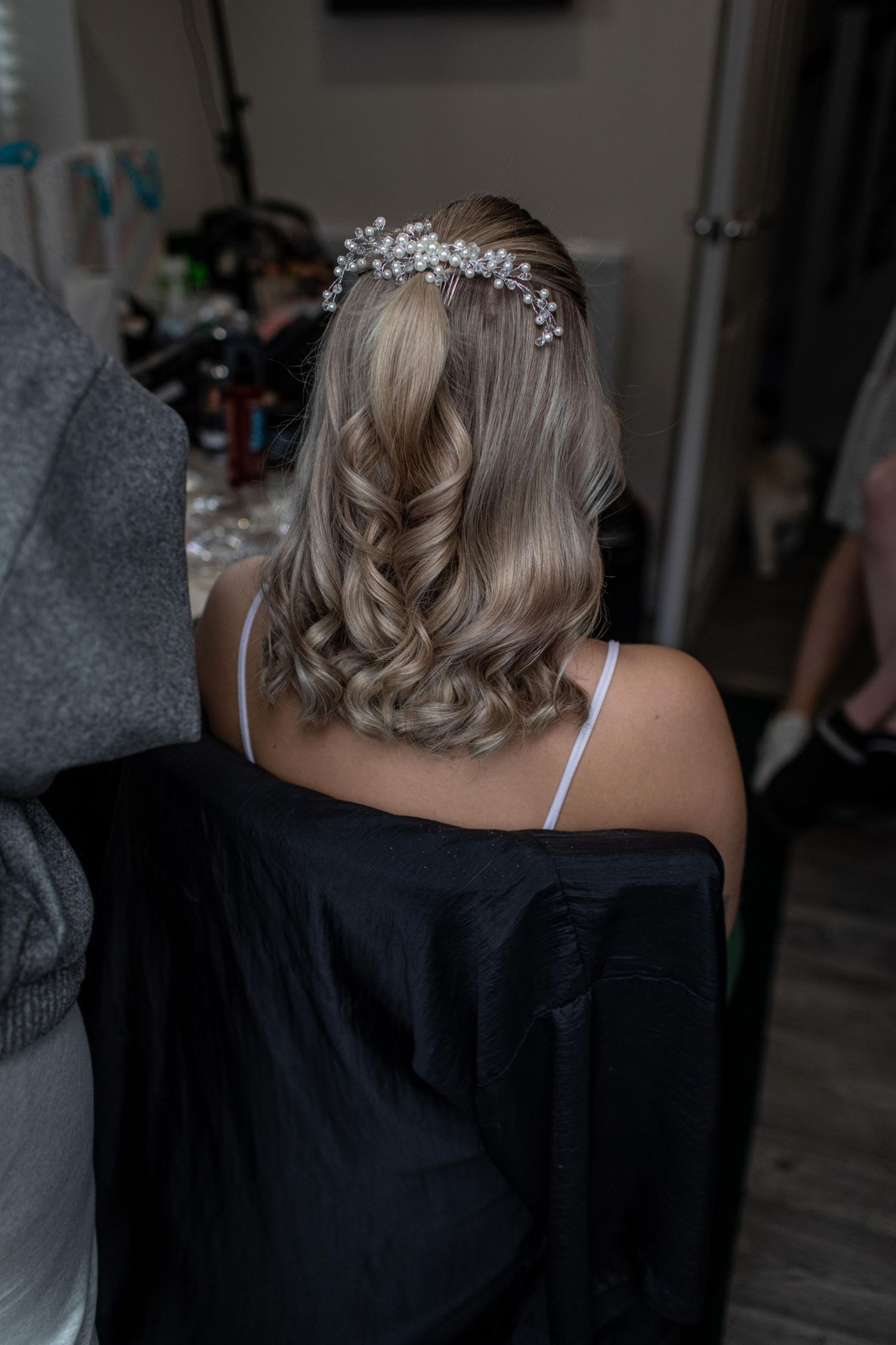 Bride with blonde curly half-up hairstyle adorned with a delicate crystal hair piece, seen from behind in a salon.