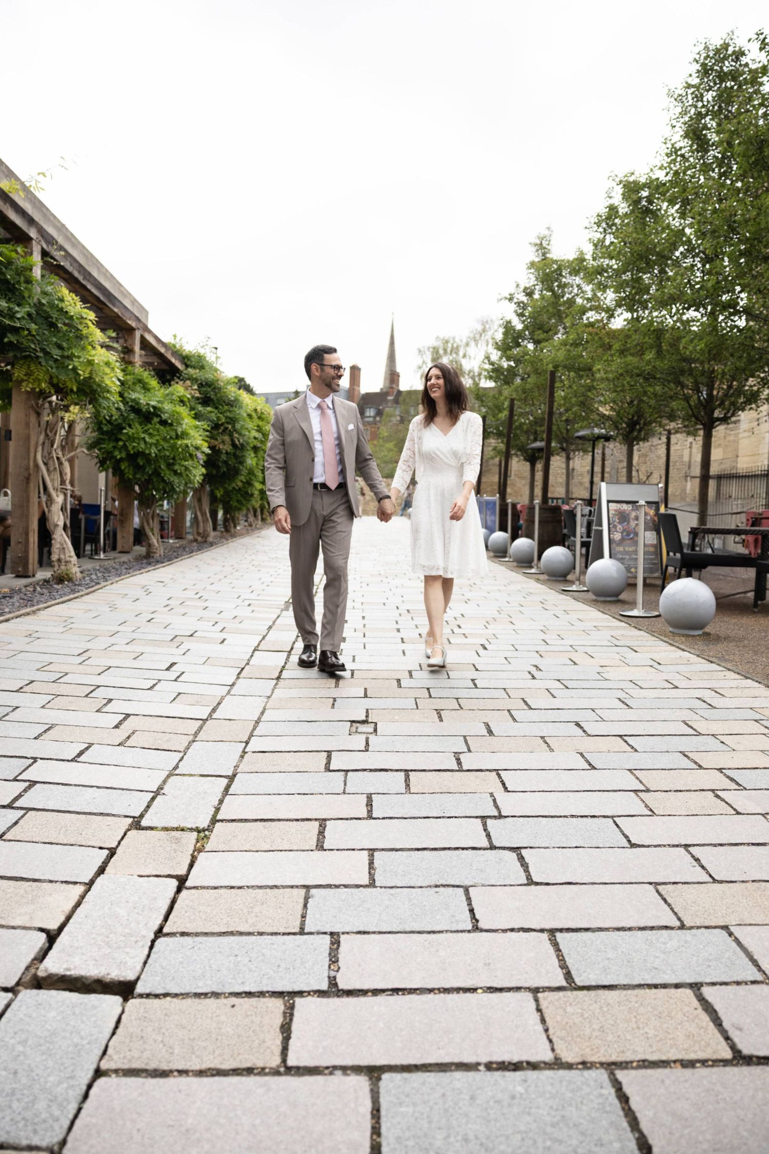 A bride and groom walk hand-in-hand along a cobblestone path lined with trees on their wedding day.