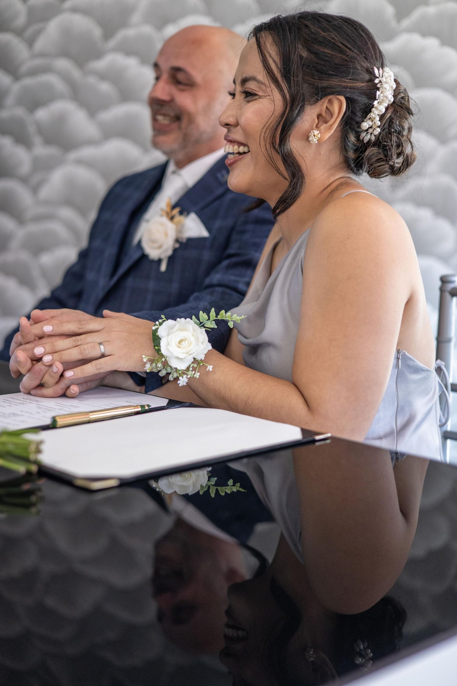 Smiling bride in gray dress with wrist corsage signs wedding documents at table with groom in blue suit behind her.