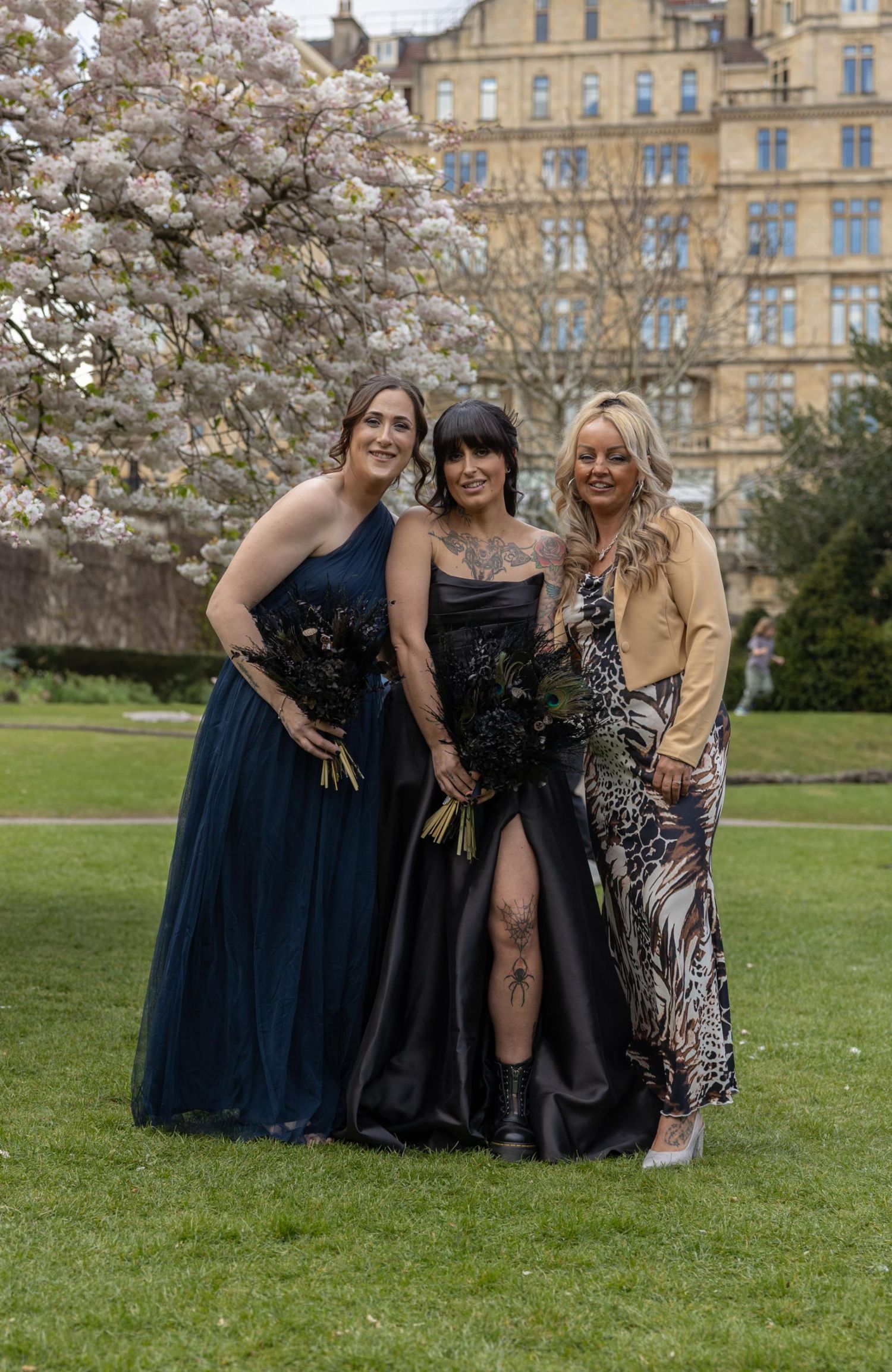 Three women pose outdoors near blooming cherry blossom tree, wearing elegant black and navy gowns at a wedding.
