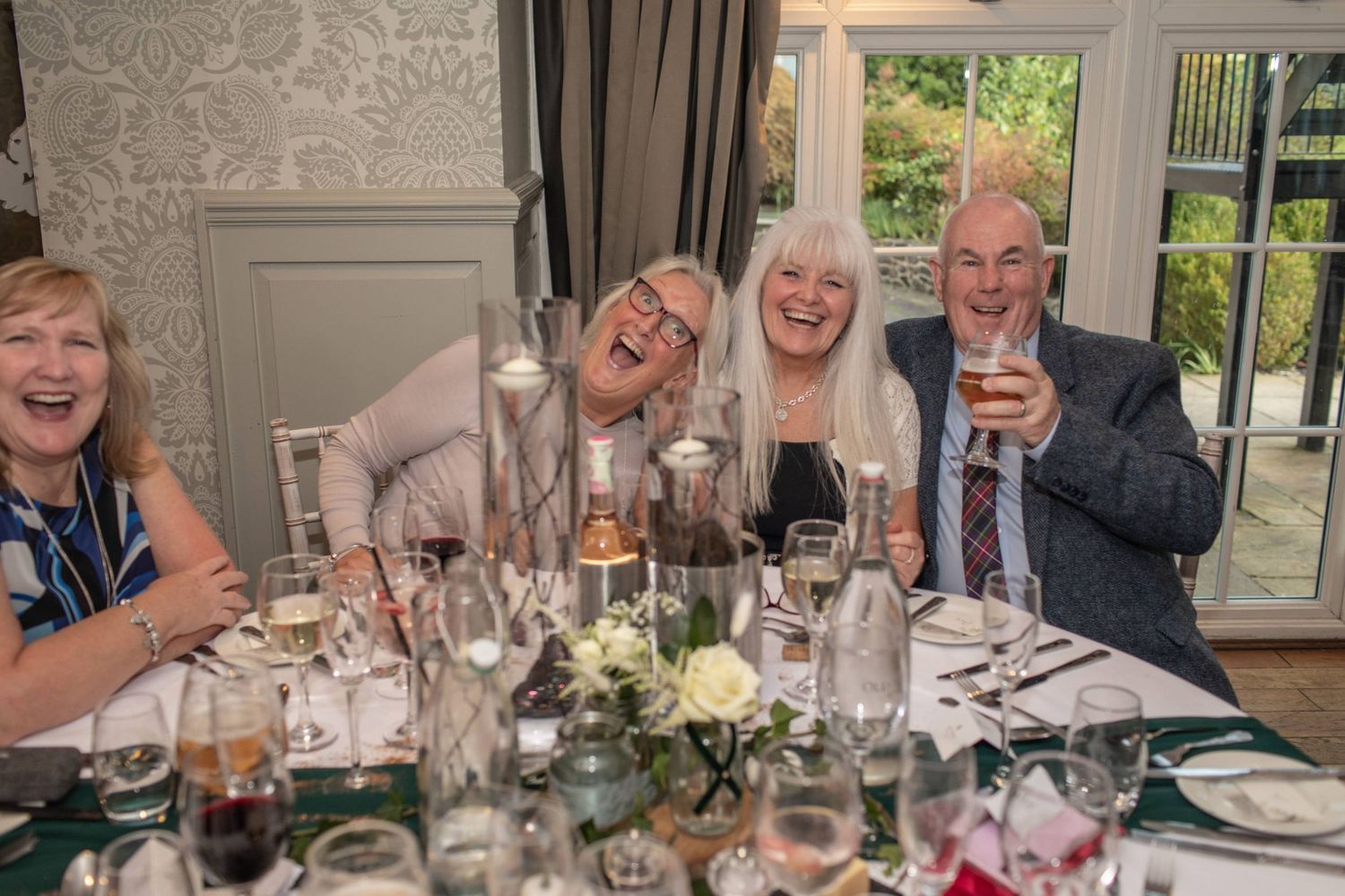 Four elderly friends laughing and raising glasses at an elegant dinner party table with floral centerpieces.