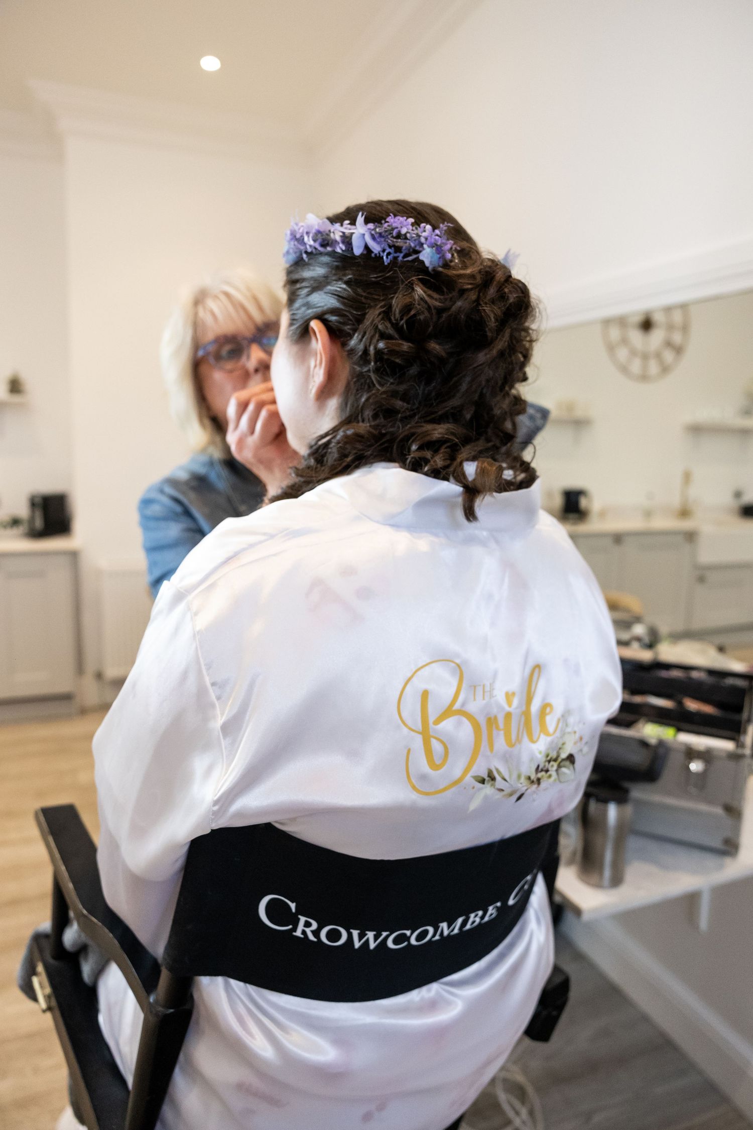 Bride in white satin robe with floral crown getting makeup done at Crowcombe Court wedding venue.