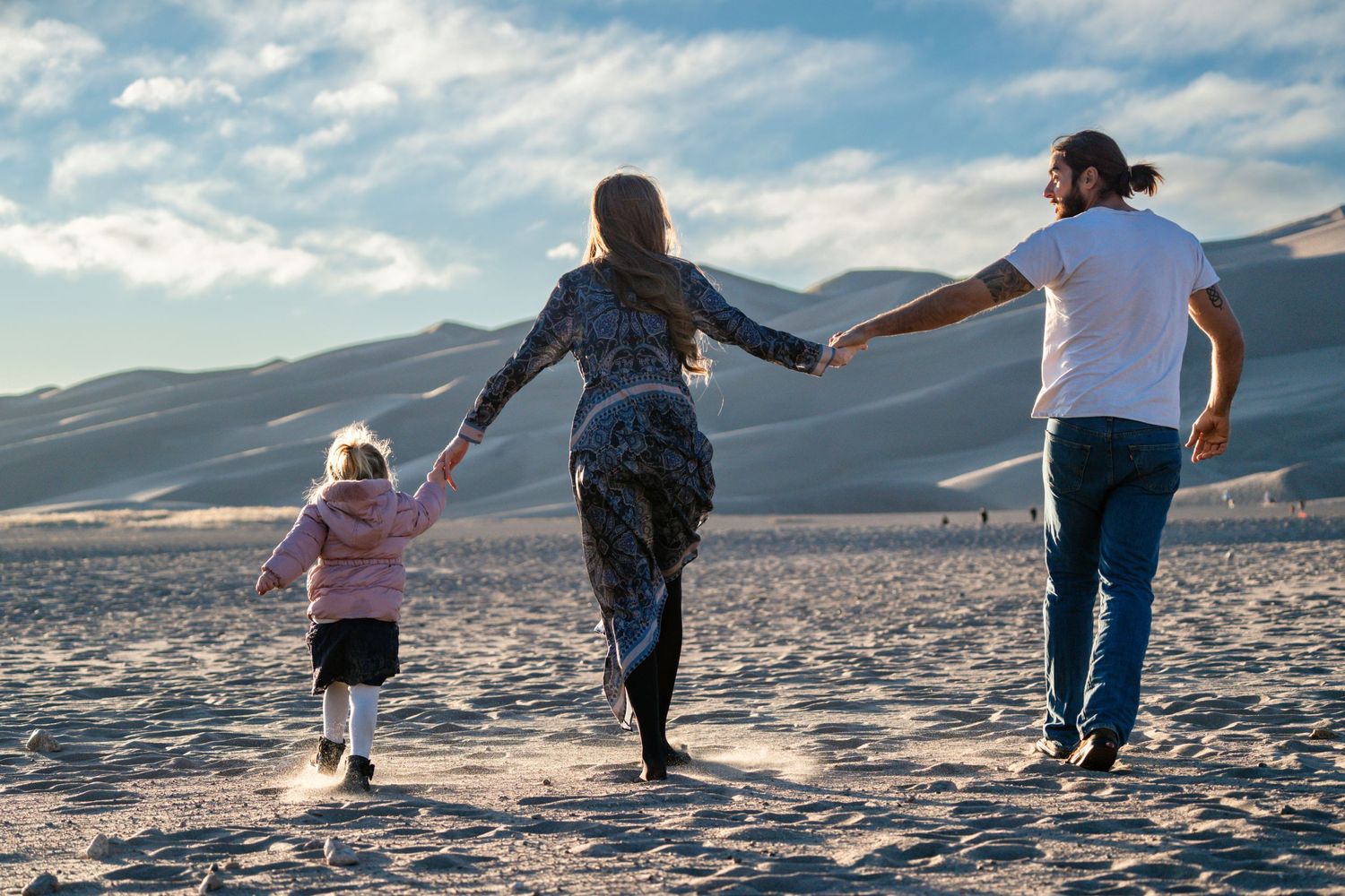 Family holds hands while walking together across sand dunes at sunset. Great Sand Dunes National Park family photographer outdoors