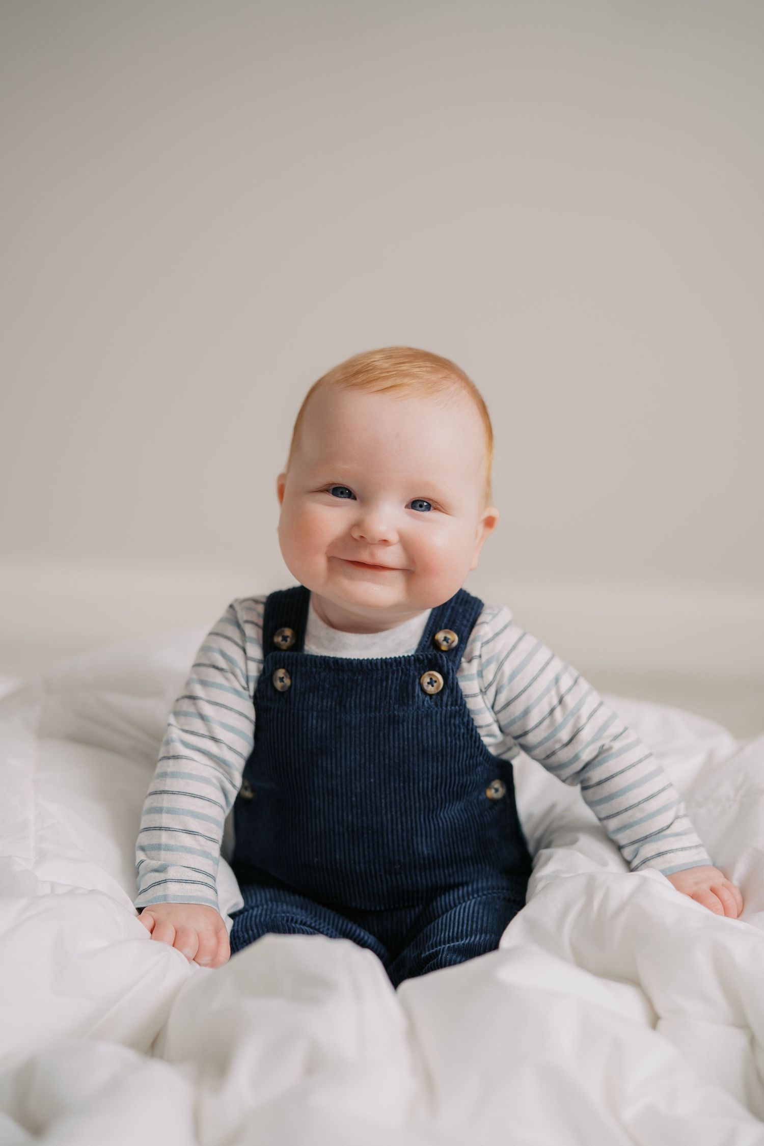 Smiling baby wearing navy overalls and striped shirt sitting on white bedding.