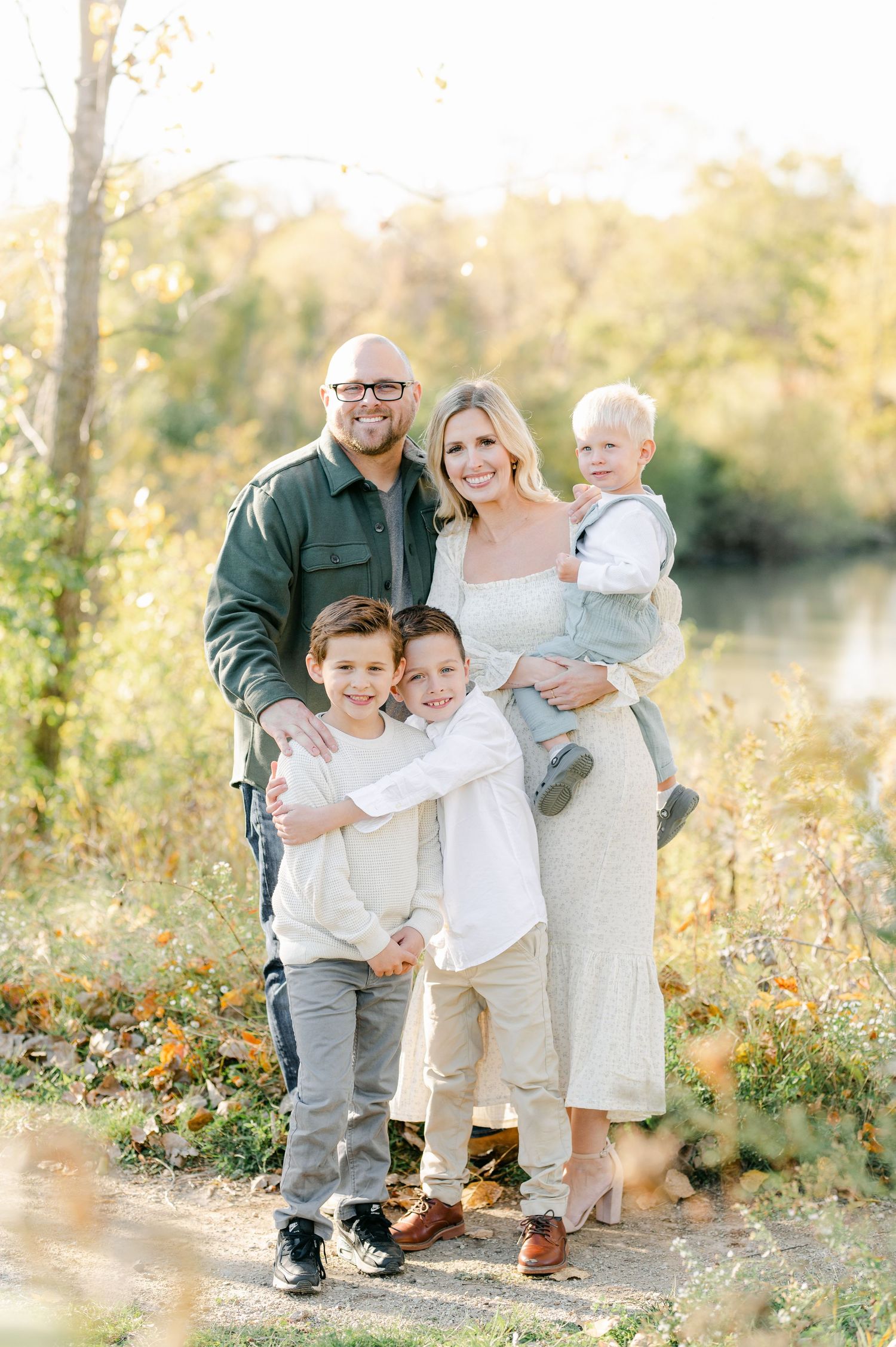 Family portrait in autumn setting with golden sunlight filtering through trees near a pond.