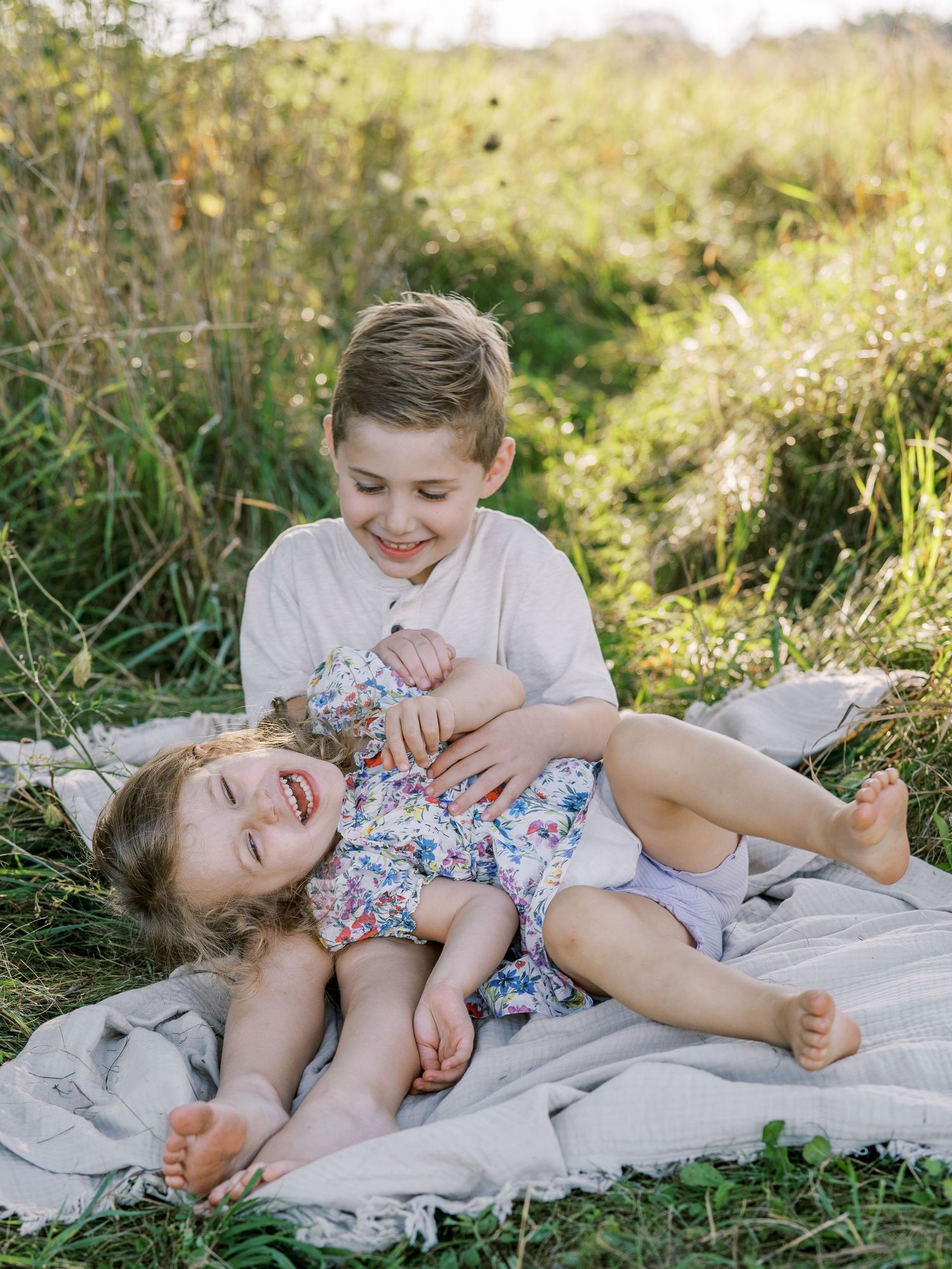 Two children playing and laughing together on a blanket in a sunlit meadow.