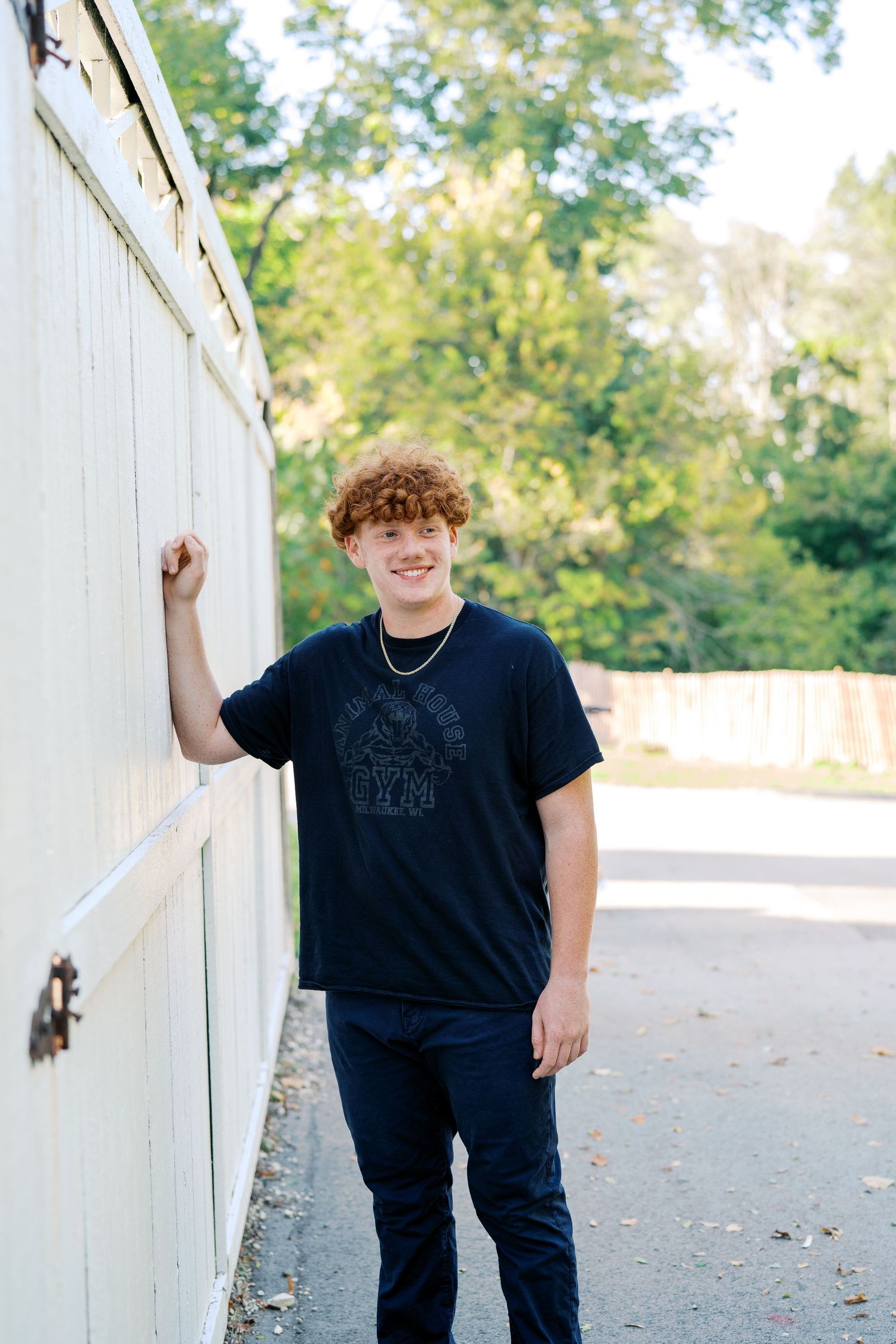 A person in a black t-shirt poses against a white fence in an outdoor setting.