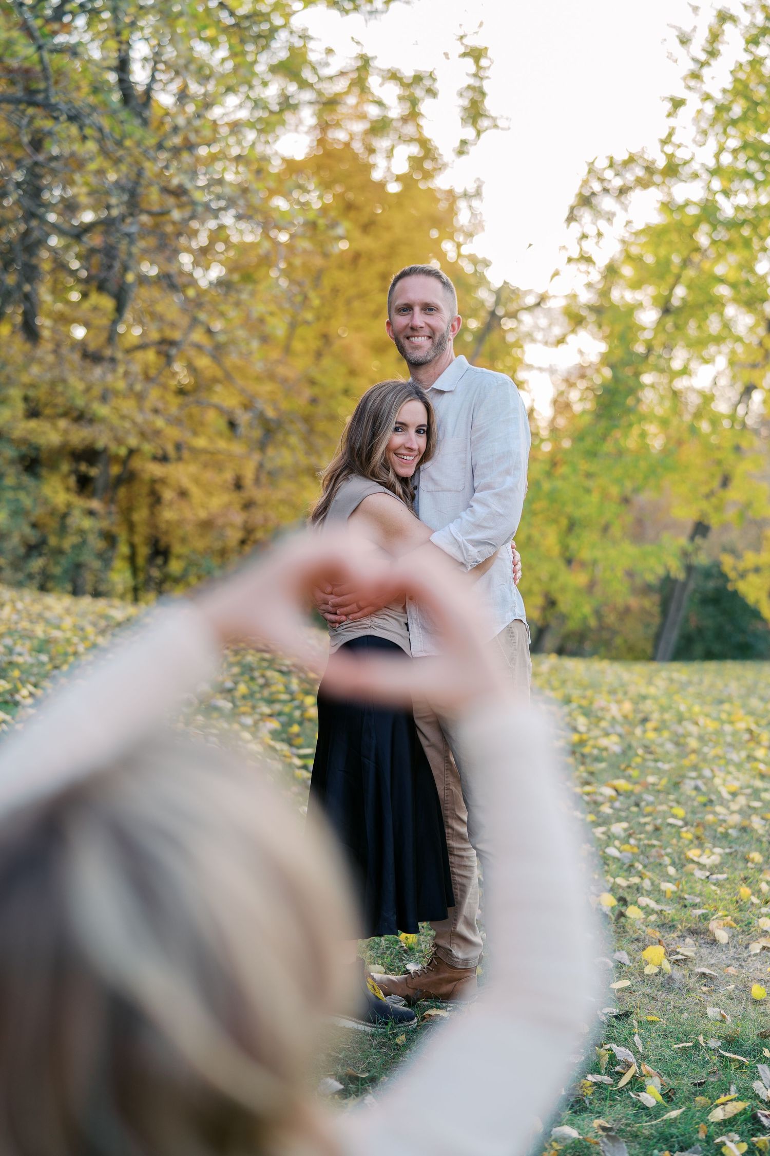 Couple poses in fall scenery while someone makes heart shape with hands in foreground.