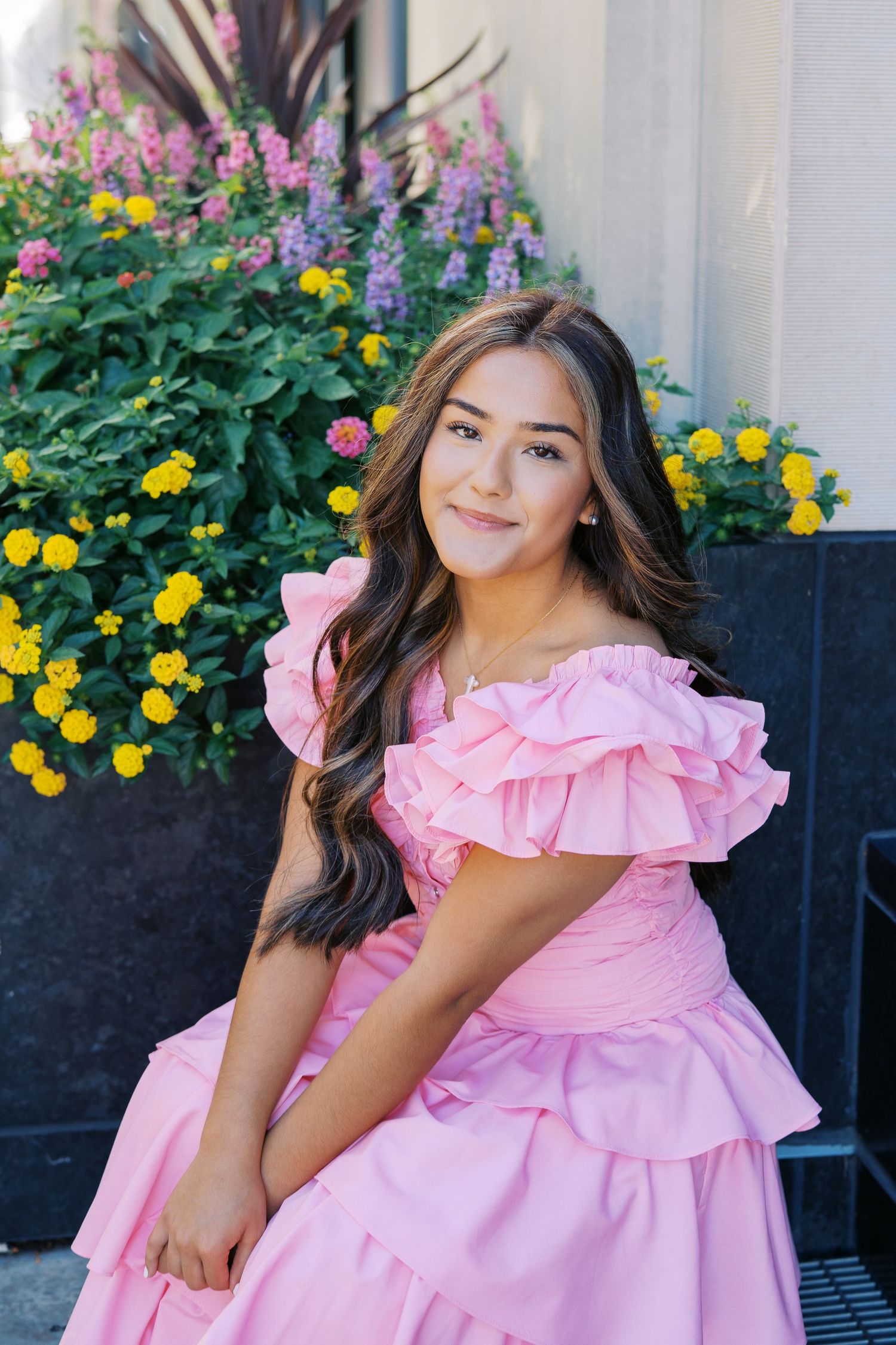 A person in a pink ruffled dress poses outdoors next to a planter filled with vibrant yellow and purple flowers.