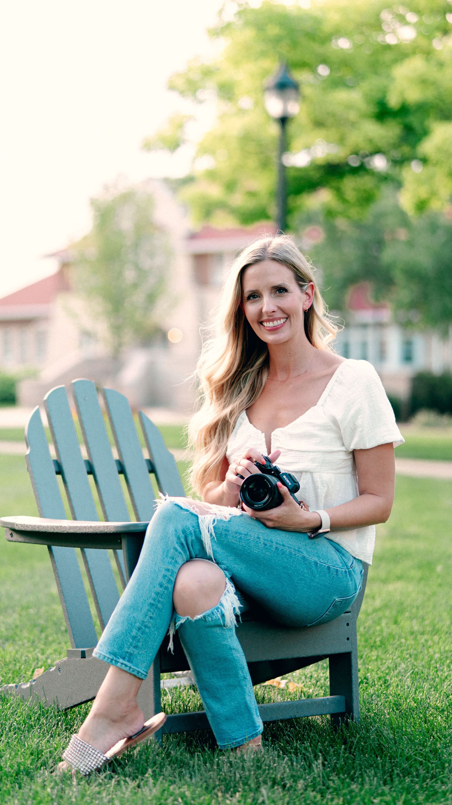 Woman in casual attire sitting on blue Adirondack chair holding camera in garden setting.