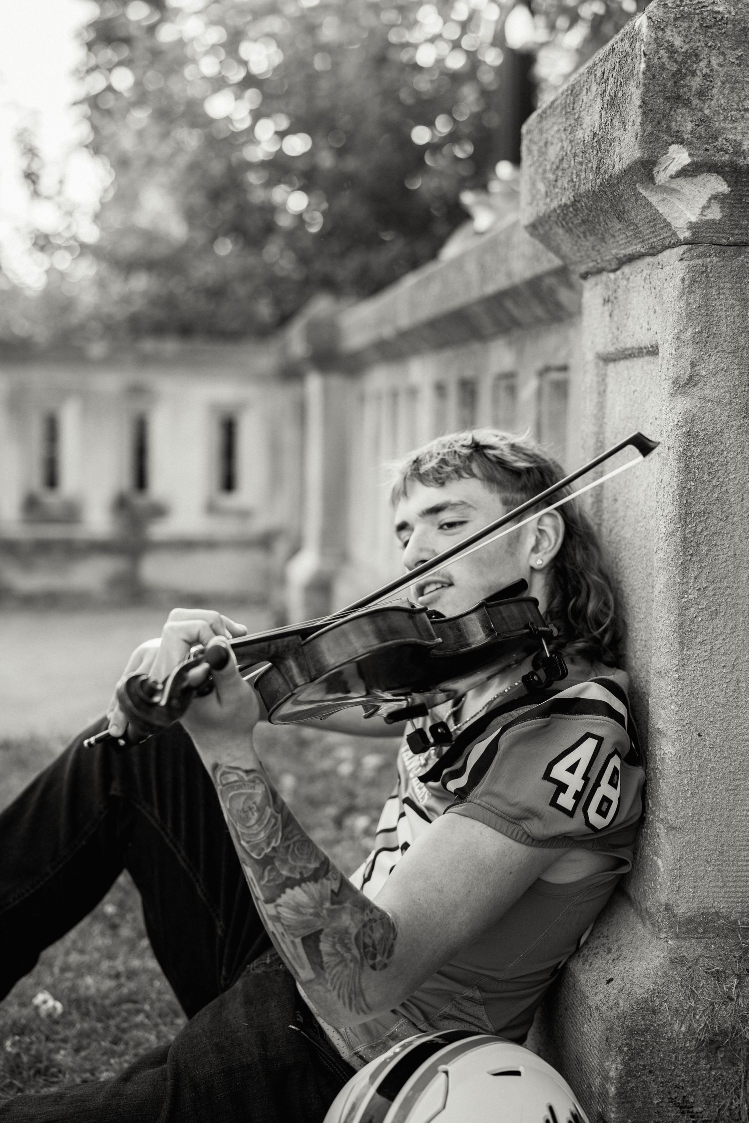 Young musician in varsity jacket playing violin while leaning against stone wall in black and white photo.