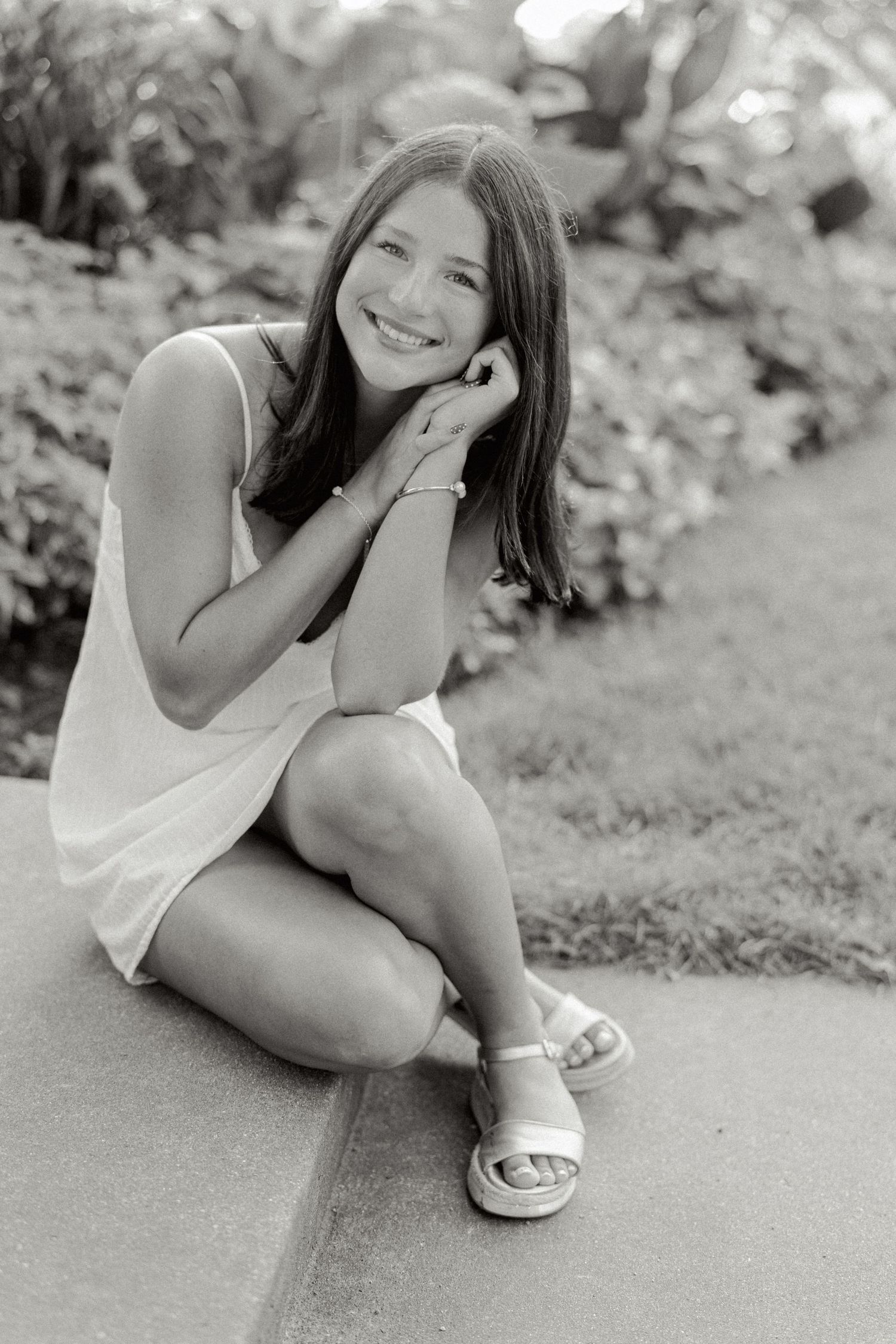 A black and white portrait of someone in a light dress sitting outdoors and smiling while resting their head on their hand.