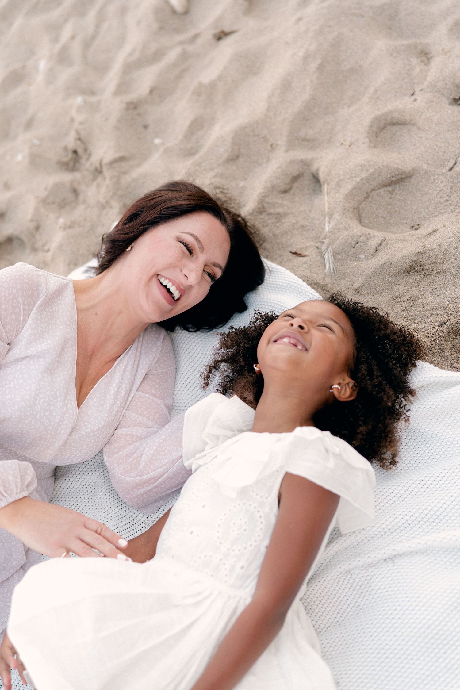 Mother and daughter sharing genuine laughter while lying on beach blanket.