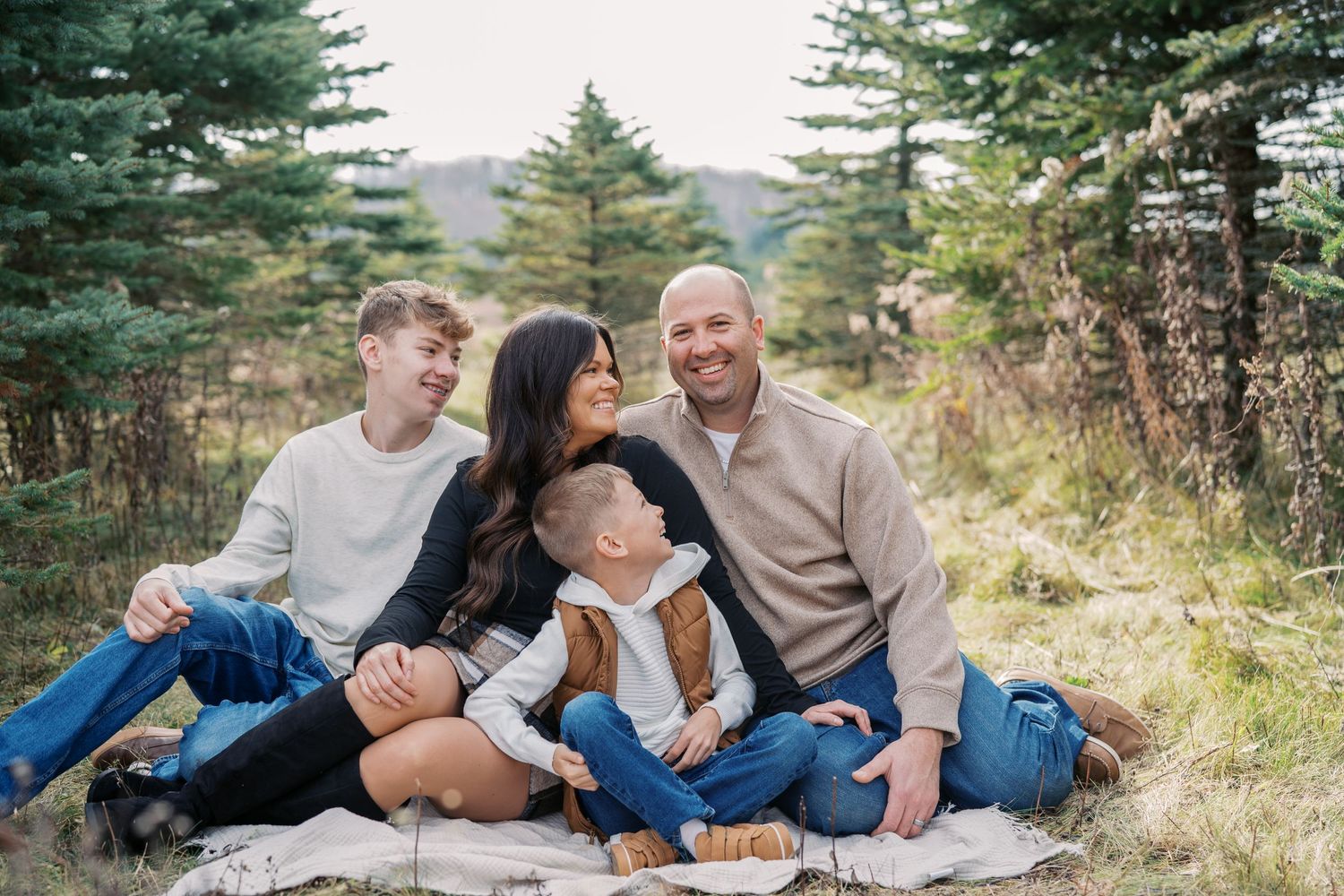 Family sitting together on a blanket in a pine forest clearing.