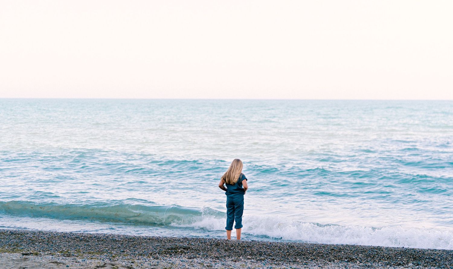 Person in blue jeans standing alone on rocky beach overlooking ocean at sunset.