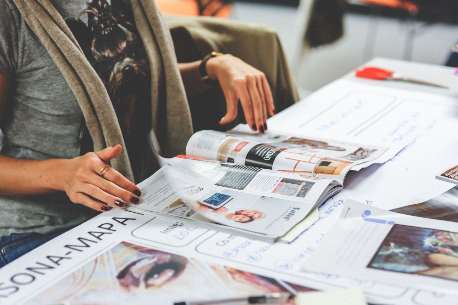 Hands reviewing magazine layouts and images spread across a wooden table surface.