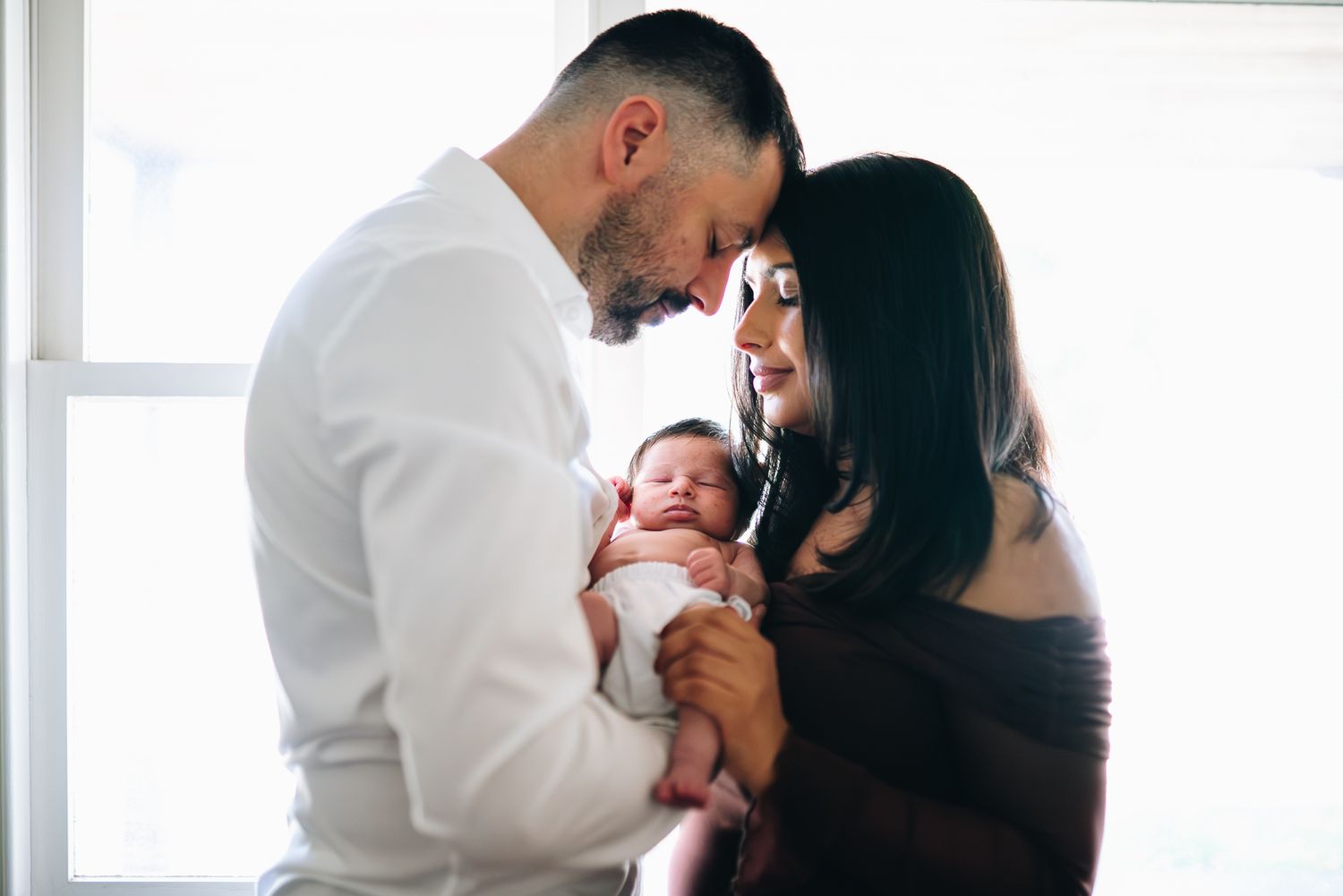 New parents share an intimate moment with their newborn baby near a bright window taken by a Melbourne newborn photographer.