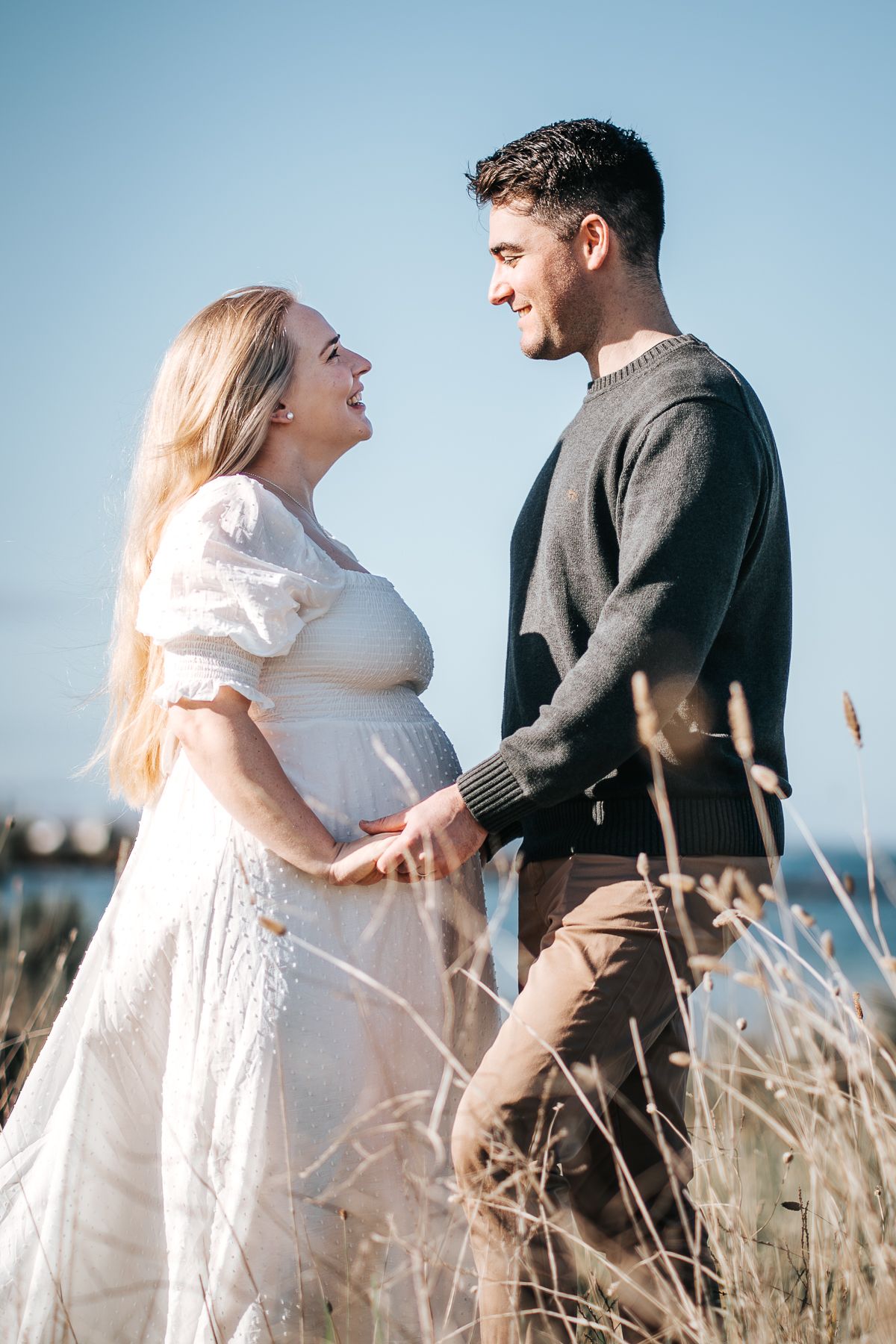 A pregnant woman in a white dress holds hands with a man in a dark sweater, smiling at each other outdoors.