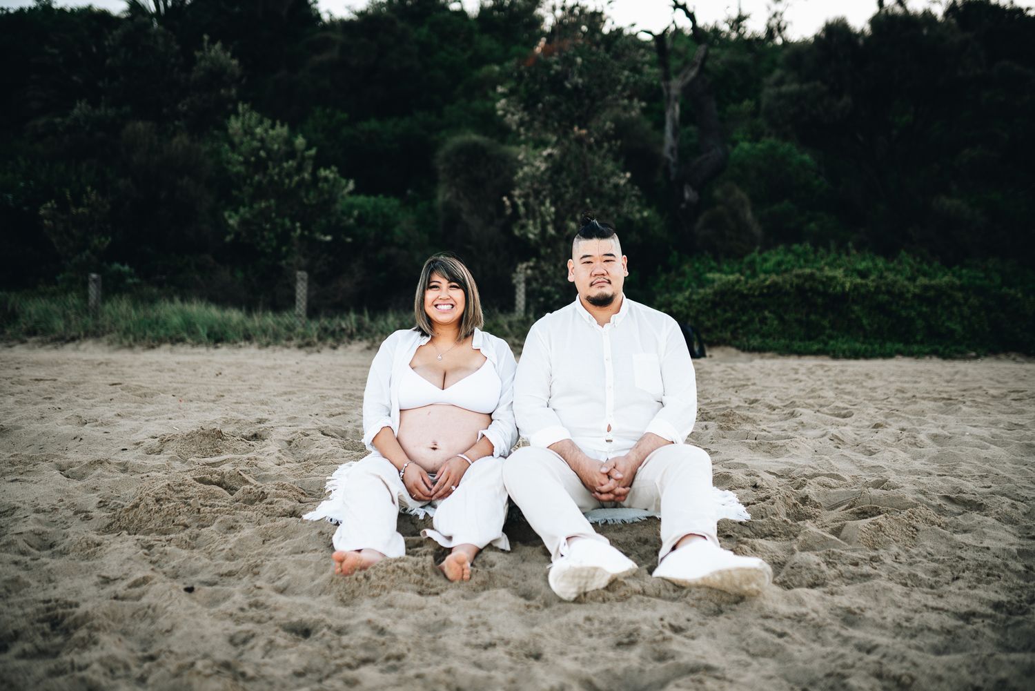 Two people in white outfits sitting together on sand with trees in the background at dusk.