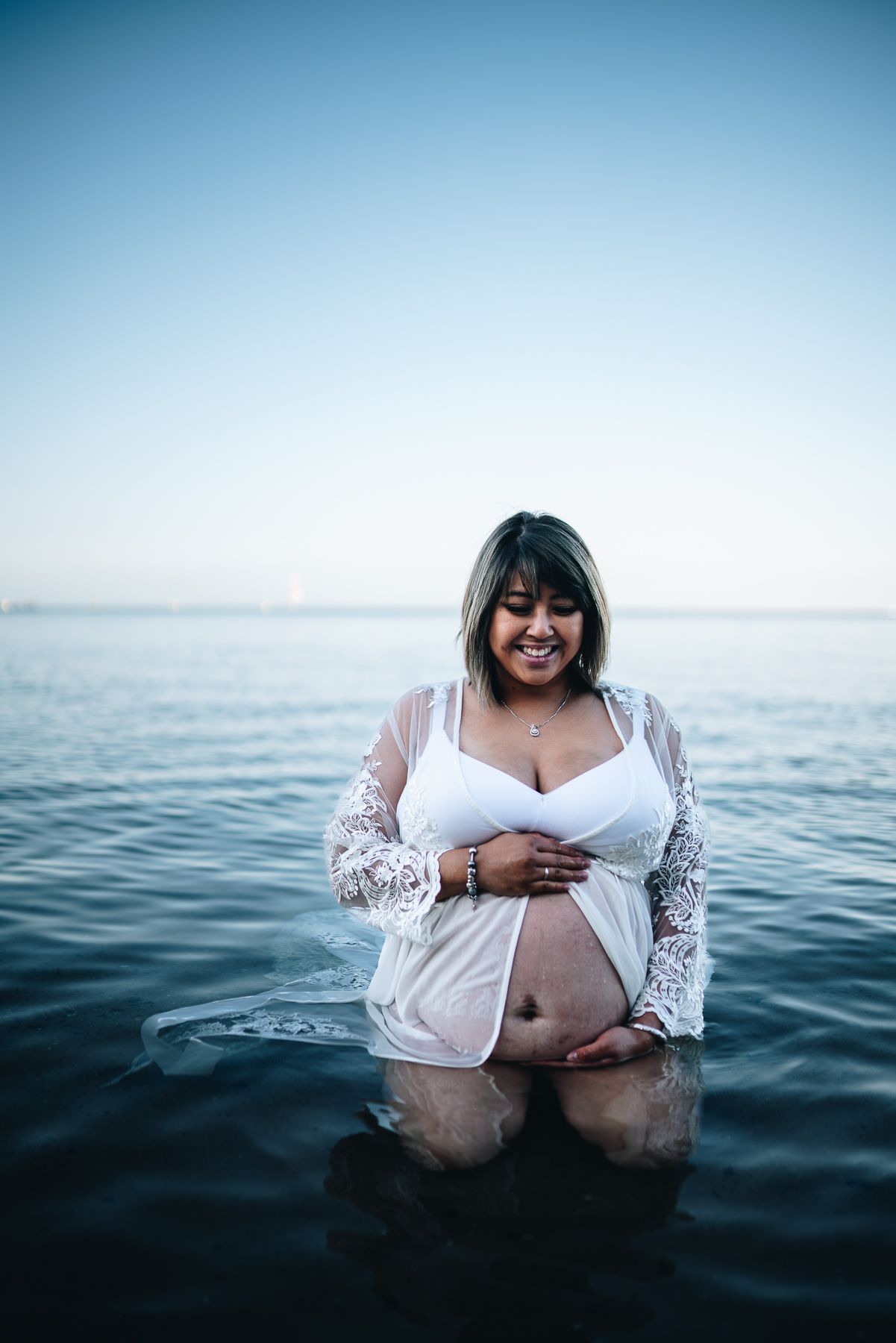 A pregnant woman in white lace stands waist-deep in calm ocean waters during a photoshoot taken by Melbourne maternity photographer.