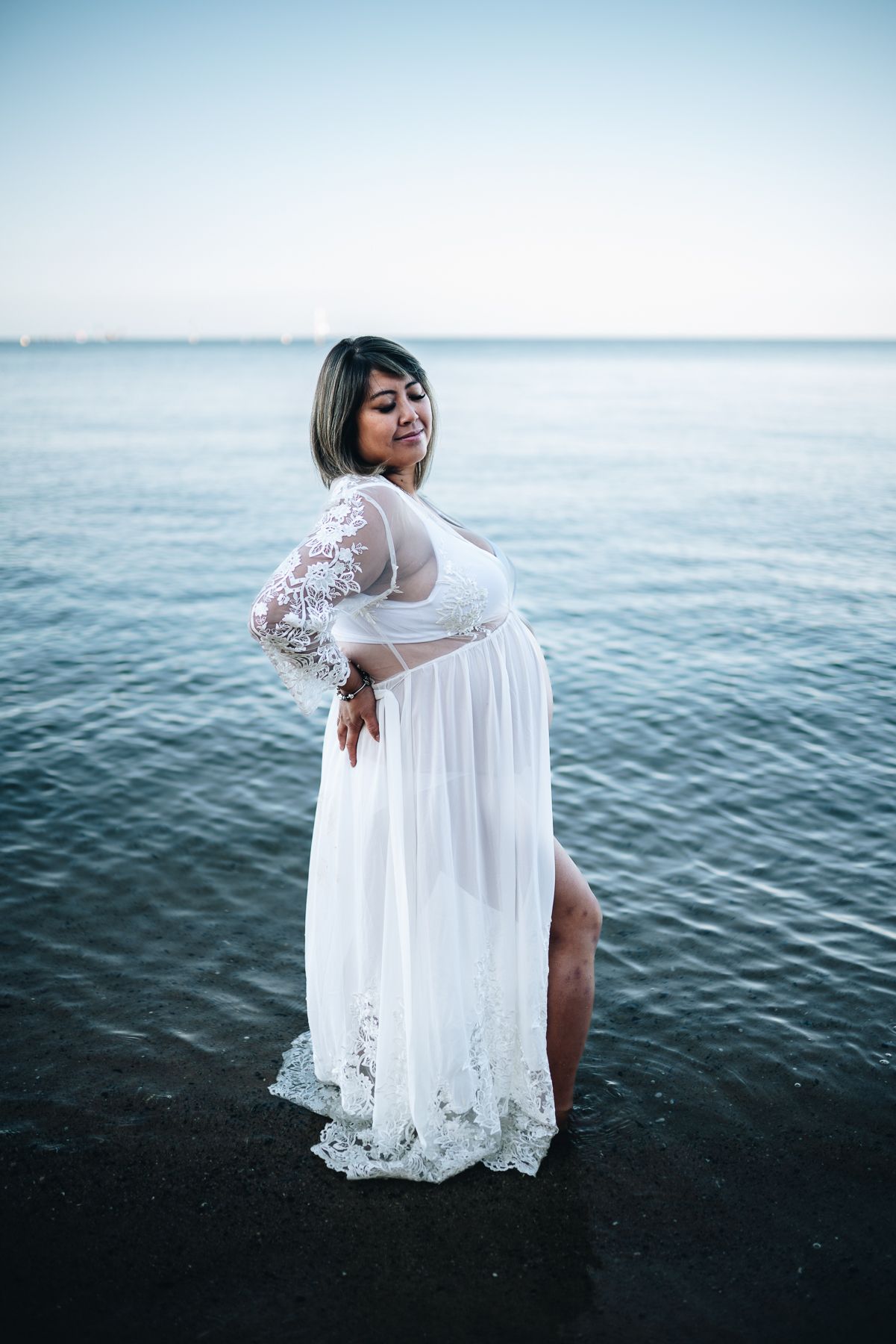 Person stands in water wearing flowing white lace dress at sunset during coastal photoshoot taken by Melbourne maternity photographer.