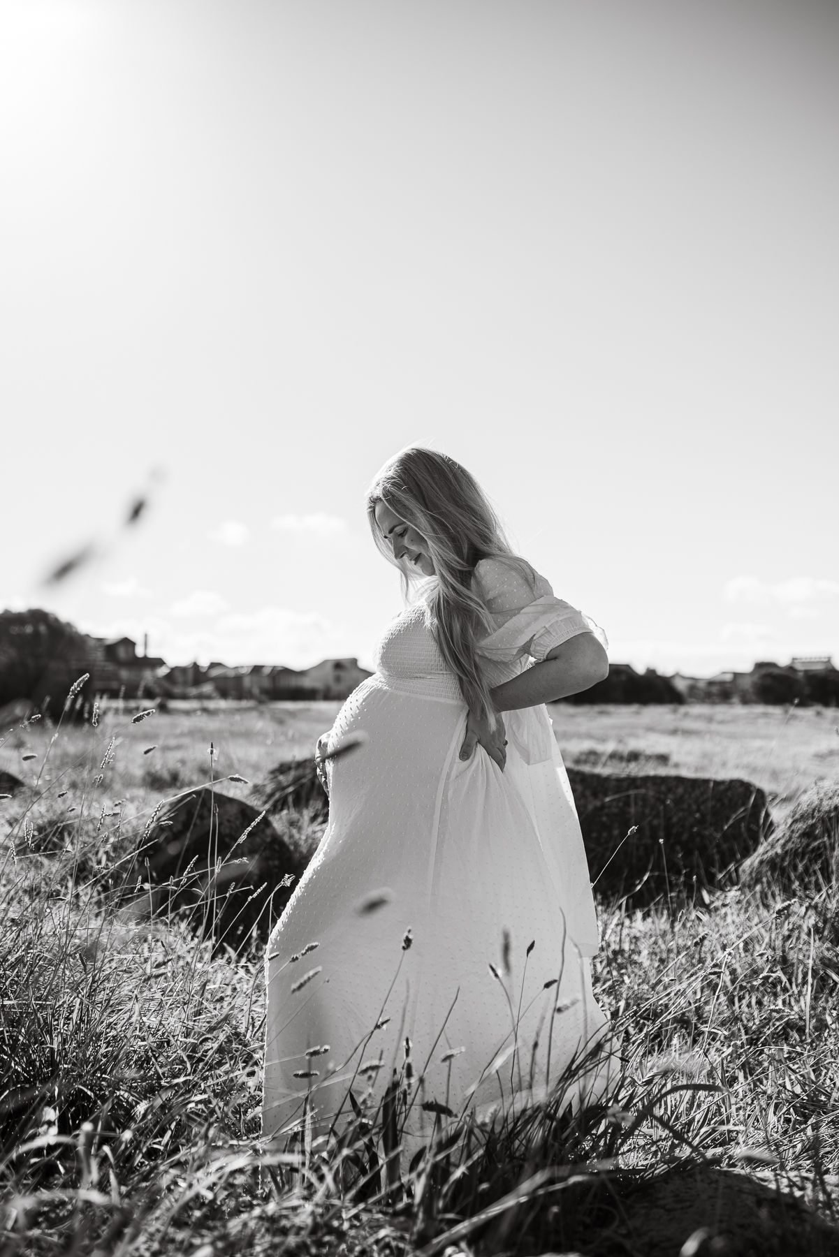 Black and white photo of a pregnant woman in a flowing white dress standing in a sunlit field.