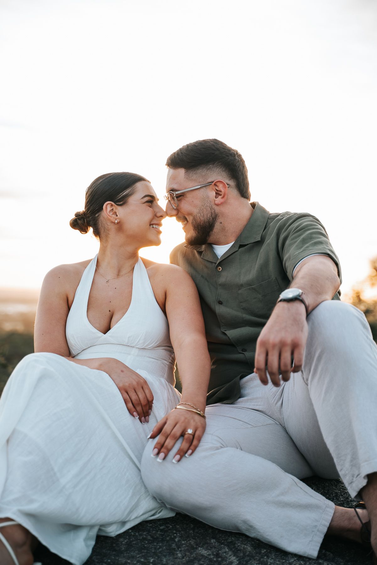 Couple sitting on rocks at sunset, smiling at each other, woman in white dress, man in green shirt taken by Melbourne couples photographer.